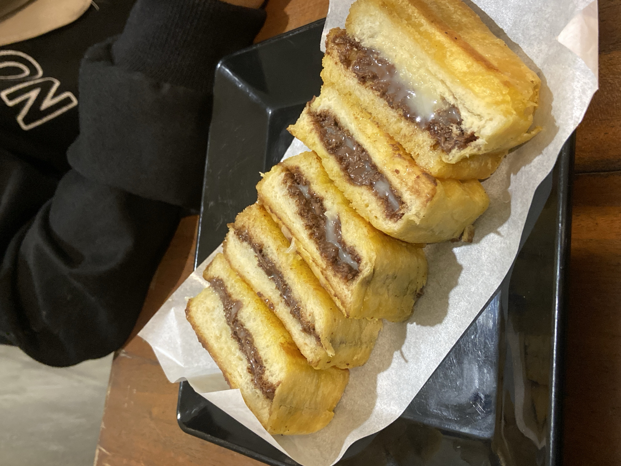 A close-up, eye-level shot captures a black rectangular plate with white parchment paper, holding five slices of a golden-brown, fried bread dessert. The bread, cut lengthwise, reveals a rich, dark brown filling, likely chocolate, with streaks of a lighter, creamy substance, possibly condensed milk. The dessert is arranged diagonally on the plate. In the background, a person wearing a black hoodie with white lettering is partially visible, with their arms resting on a wooden table. The setting appears to be a casual eatery.