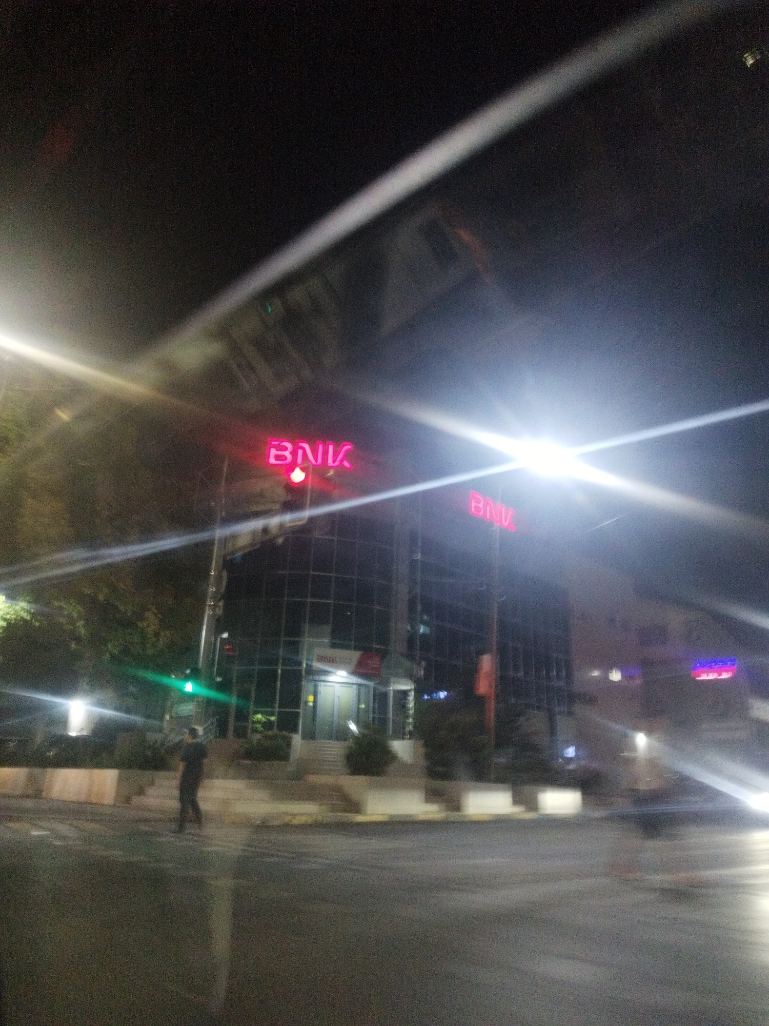 The image depicts a street scene at night in Shymkent, Kazakhstan. A modern, glass-fronted building with a red neon sign reading "BNK" is prominently featured. To the left of the building, a traffic light with a green signal is visible. A lone figure, dressed in dark clothing, stands on the sidewalk in front of the building. The street in the foreground is wet and reflects the surrounding lights. The overall atmosphere is urban and nocturnal, with significant light glare from streetlights and possibly vehicle headlights.