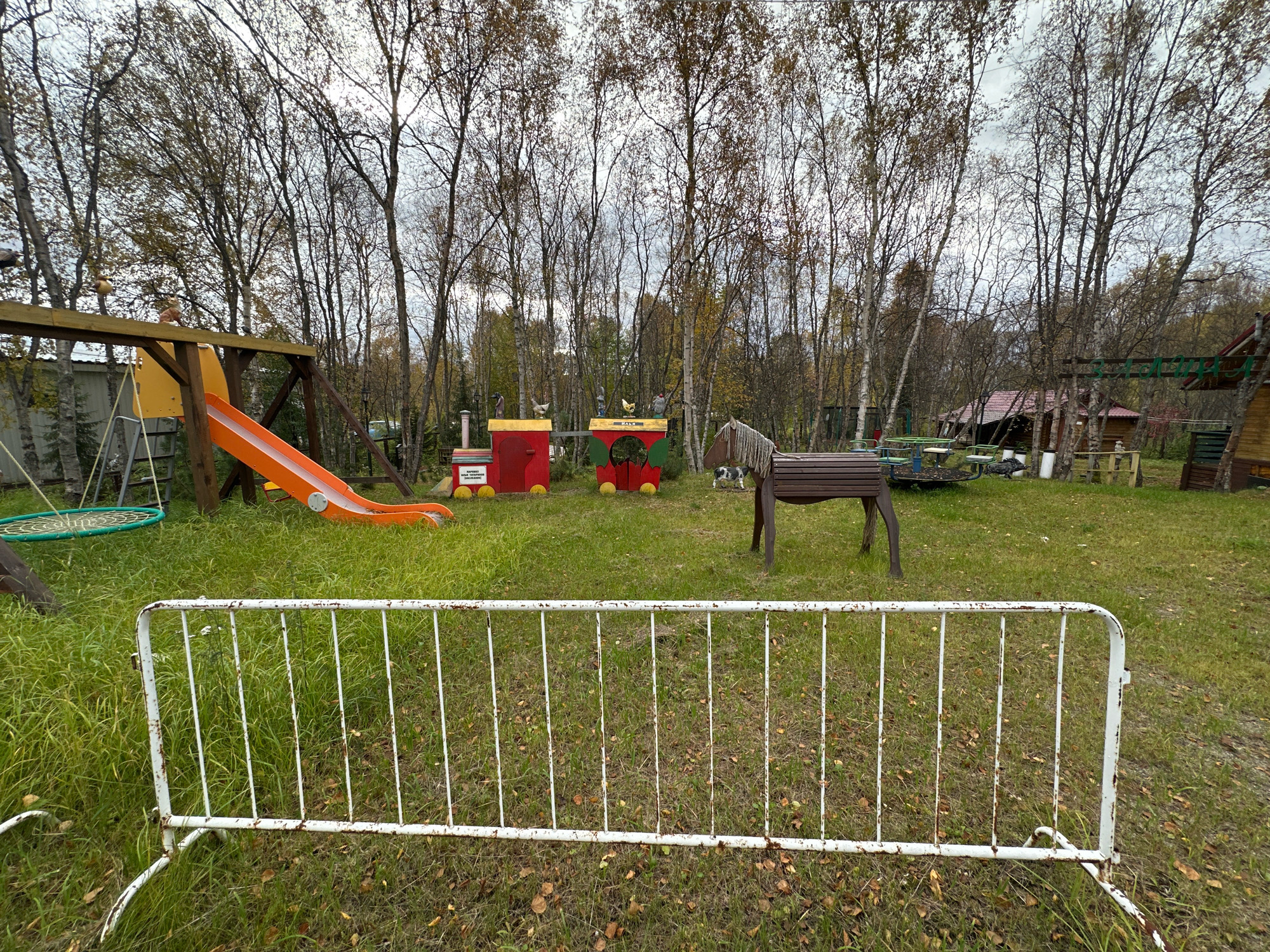 An outdoor, daytime, wide-angle shot shows a children's playground. In the foreground, a white, rusted metal barrier runs horizontally across the frame. Behind the barrier, a grassy area with some fallen leaves is visible. To the left, a wooden swing set with an orange slide and a green disc swing is present. In the center of the playground, there are toy train cars made of red and green wood. A wooden horse with a brown saddle and white mane stands to the right of the train cars. In the background, a dense forest of deciduous trees with sparse foliage and an overcast sky can be seen. A few rustic wooden structures are partially visible among the trees. The scene appears to be located in Zverosovkhoz, Russia. There are no people visible in the image.