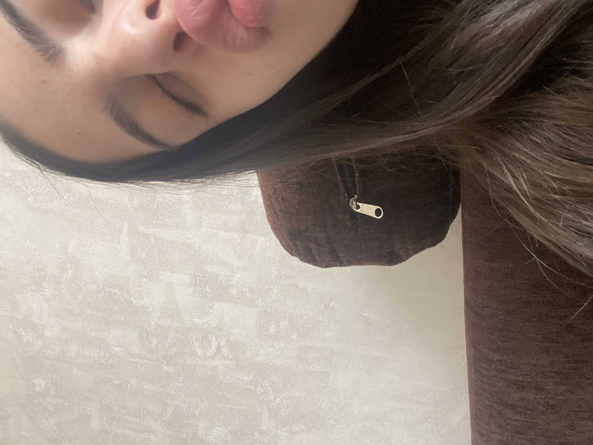 Here is a forensic description of the image:
The image shows a close-up, rotated view of a person's face, specifically the nose, lips, and closed eye. The person has dark hair. They are wearing a brown garment with a zipper. The background appears to be a light-colored wall.
There is no information available in the image to determine the location in Dragovishtitsa, Bulgaria.