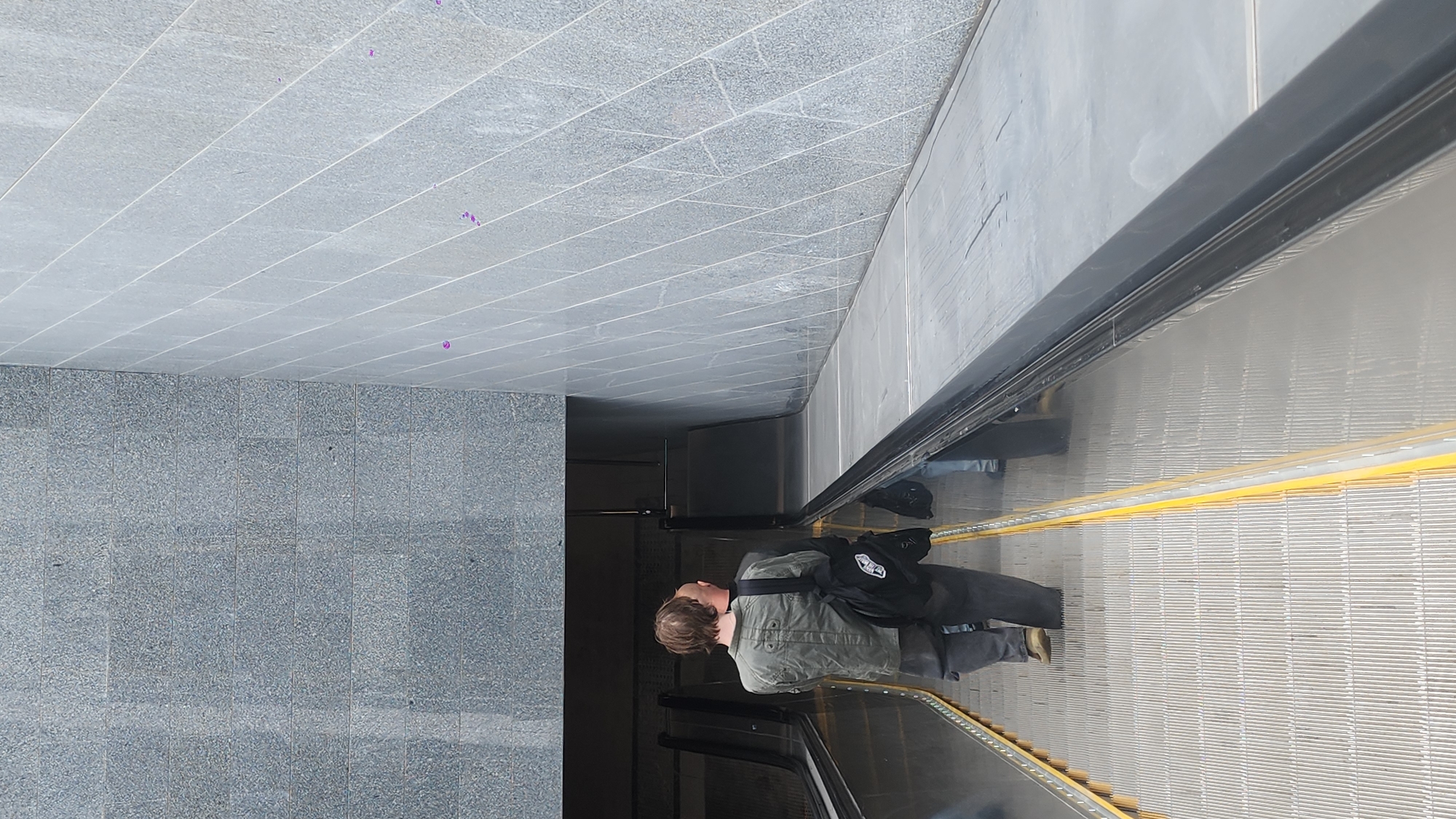 A man with fair hair and wearing a green jacket, dark pants, and tan shoes rides an escalator in what appears to be a subway station in Moscow, Russia. He carries a black backpack over one shoulder. The escalator is flanked by gray tiled walls and a matching tiled ceiling. The escalator steps are metal with yellow safety markers. The lighting appears to be natural, coming from the station's entrance. The image appears to be taken from above.