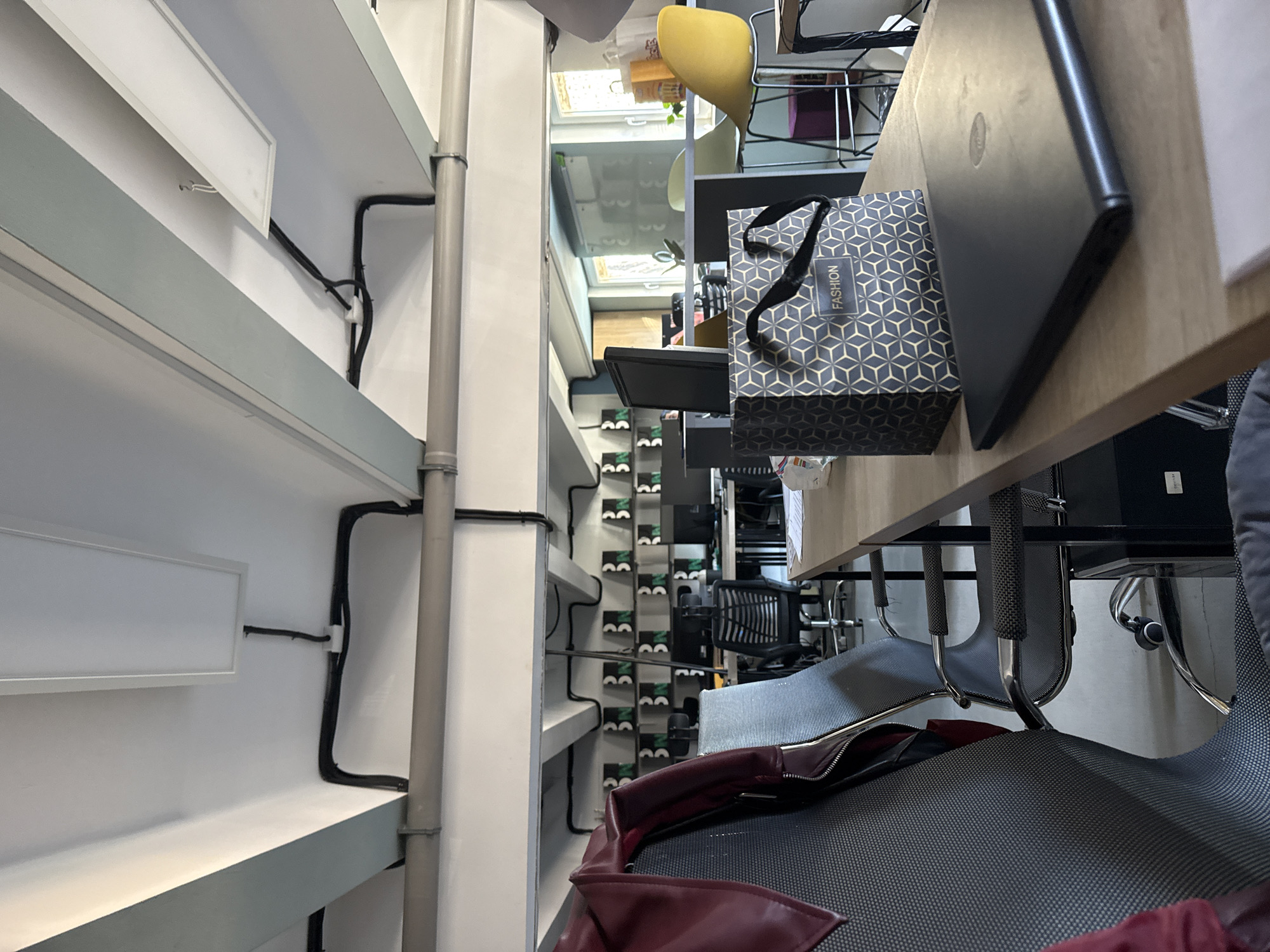 This is an indoor shot of a desk area, though the angle makes spatial orientation difficult. A desk surface is visible, with a black laptop and a blue geometric patterned gift bag on top. Below the desk, there's a gray office chair with a red garment draped on it. To the left of the desk area, there's a wall with shelves, and lighting fixtures are mounted on the shelves with some electrical cables running along the wall. The vertical pipes run along the side of the shelving. The location and city are not identifiable from the image.