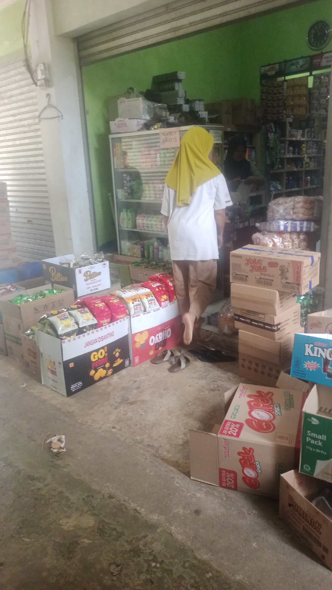 The image depicts an outdoor market stall, likely in Cogreg, Indonesia, featuring a display of packaged snacks and goods. A person wearing a yellow hijab and a white shirt stands with their back to the camera, facing a brightly lit shop interior. The stall is cluttered with cardboard boxes of various sizes, many containing snack products. Prominently displayed are boxes of "GO! PRIME" chips and "OKHHO" snacks. To the right, stacked boxes include "Yale Yale" snacks and a blue box labeled "KING." Further into the shop, shelves are stocked with more goods. The ground is a concrete surface, showing some wear and debris. Two pairs of sandals are visible on the ground near the snack boxes. The overall scene suggests a retail environment with merchandise laid out for sale.