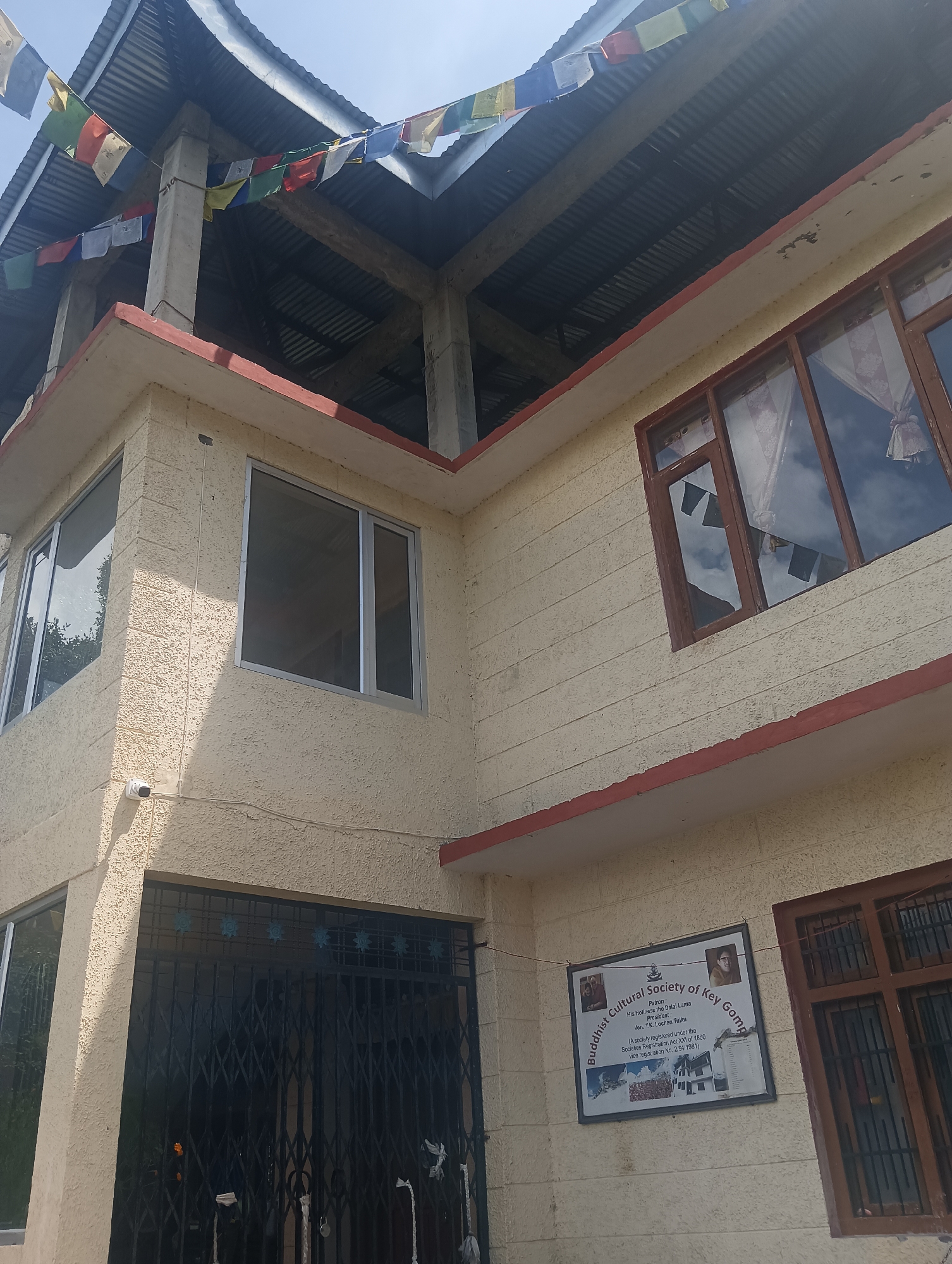The image shows a two-story building with a textured, light-colored exterior. Prayer flags are strung across the top of the structure, which appears to be under construction or renovation, with exposed beams and corrugated metal roofing visible. The building has several windows, some with wooden frames and others with metal frames. There is a black metal gate on the ground floor. A sign on the wall reads "Buddhist Cultural Society of Key Gompa" and lists His Holiness the Dalai Lama as Patron and Ven. T.K. Lochen Tultu as President. There are two small images of individuals on the sign. A security camera is mounted on the exterior wall near the upper corner of the ground floor. The scene is outdoors, under a bright sky.