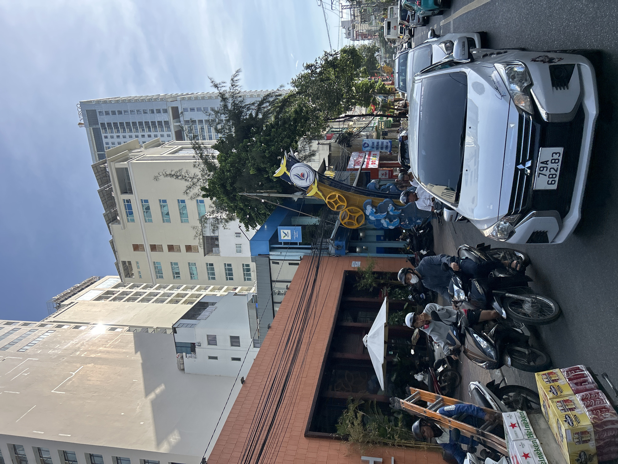 The image depicts a street scene in Nha Trang Ward, Vietnam. The street is lined with tall buildings, including hotels and apartments. The foreground shows several motorbikes and cars, indicating traffic on the road. One motorbike carries two individuals wearing helmets. Another motorbike has a person in the rider's seat and a ladder strapped to the side. A white car with a license plate reading "79A 682.83" is prominently featured. Boxes of "Heineken" and "Strongbow" are visible near the front of the frame. The sky is clear and blue. The architecture and the presence of motorbikes suggest a bustling urban environment typical of Vietnam. A large, colorful art installation or signage is visible on the right side of the street, featuring a circular logo with waves and bird-like elements.
