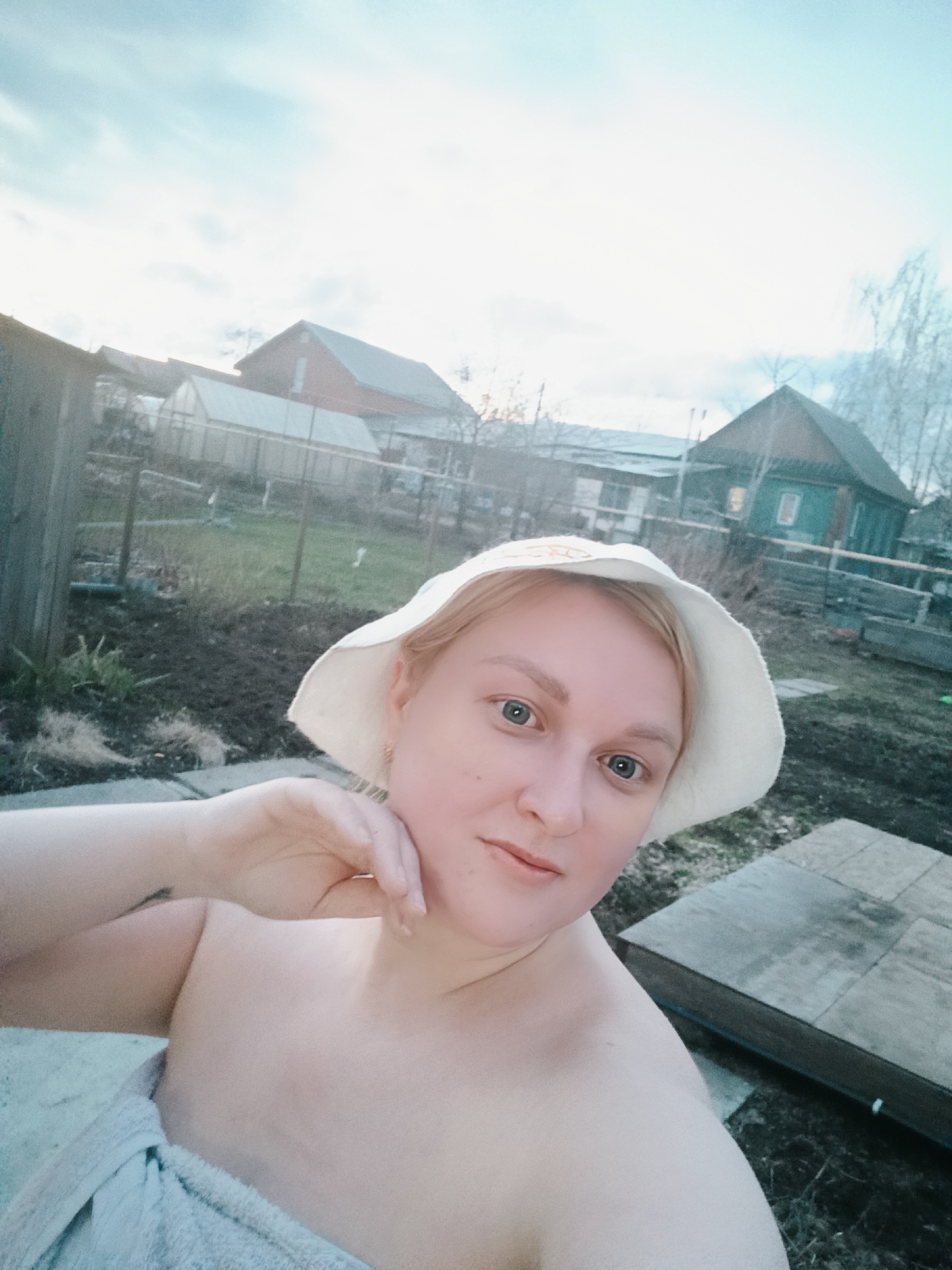 The image depicts a woman in a white hat and a towel, standing outdoors. Her left hand is raised to her chin. In the background, there are several buildings, including a greenhouse, a red barn-like structure, and a green wooden house with a light on inside. The scene appears to be in a rural or semi-rural area with vegetation and overcast skies. The location is specified as being in the city of Chaikovsky, Russia.