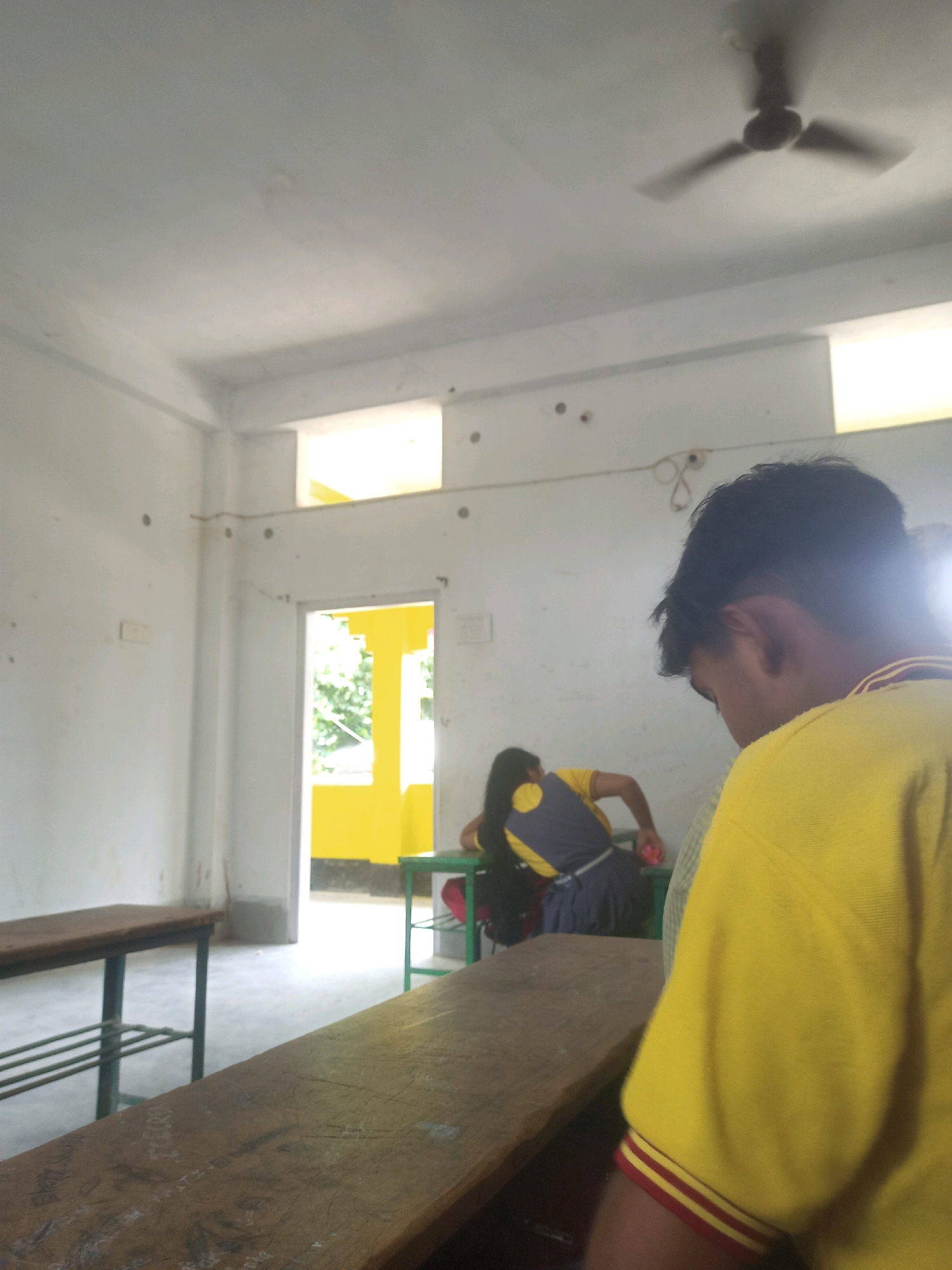 This is a medium-angle, indoor shot of a classroom in Hatsinigmari, India. The walls are painted white, and the ceiling is also white with a ceiling fan visible. There are two windows in the room, one on the right and another above the doorway on the back wall, both letting in natural light. A doorway on the back wall leads to an area painted bright yellow. In the foreground, a wooden desk is partially visible, with scratches and inscriptions on its surface. To the left, a wooden bench with a metal frame is seen. In the mid-ground, a person wearing a yellow shirt is visible from the back, looking down. Further back, another person, possibly a student, is seated at a table, wearing a blue and yellow uniform and facing away from the camera. The floor is light-colored concrete.