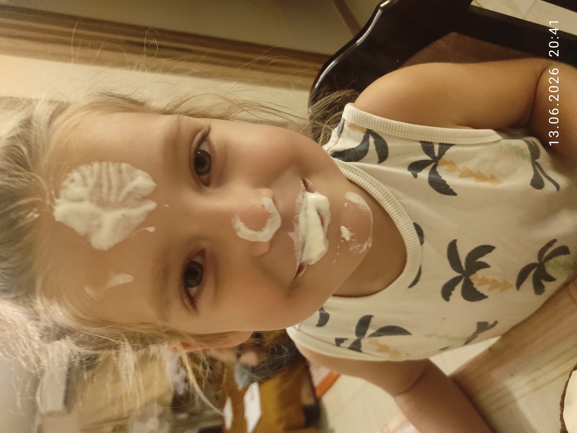 A young girl with blond hair, her face smeared with white frosting, smiles at the camera. She is wearing a white tank top with a palm tree pattern. The background suggests an indoor setting, possibly a home, with blurred furniture and a doorway visible. The timestamp "13.06.2026 20:41" is overlaid on the bottom right of the image, indicating the date and time the photograph was taken. The image does not contain any information about the specific location in Mrčevac, Montenegro.