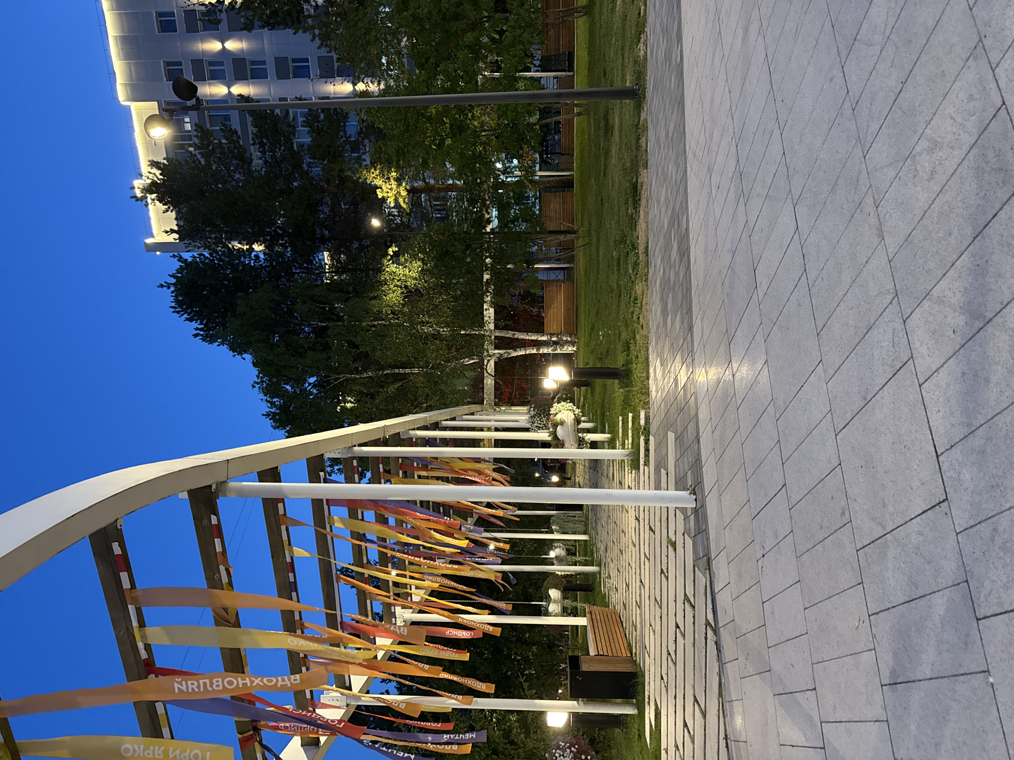 The image depicts an outdoor public space in Noyabrsk, Russia, at dusk. A white, arching pergola is prominently featured, decorated with numerous colorful ribbons hanging from its beams. The ribbons are inscribed with text in Cyrillic, suggesting a festive or commemorative purpose. A paved walkway, constructed with large, rectangular light-grey tiles, extends into the distance beneath the pergola. To the left of the walkway, there is a wooden bench and a dark trash receptacle.
Beyond the pergola, a well-maintained grassy area is visible, dotted with trees. In the background, a multi-story building with illuminated windows and decorative lighting along its facade stands tall. A streetlamp with a distinctive modern design is positioned in front of the building. The overall scene suggests a park or a public square designed for relaxation and events, illuminated by ambient lighting as daylight fades. The sky is a deep blue, characteristic of twilight. There are no people visible in the frame.