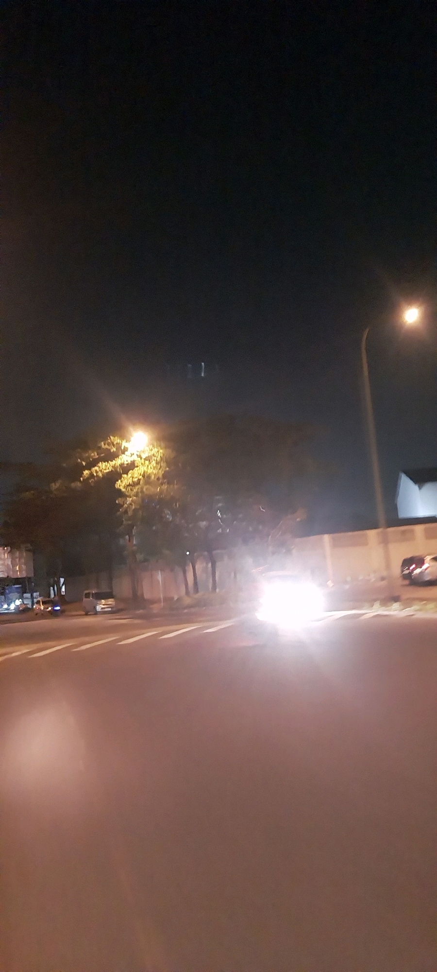 This is a nighttime, slightly blurred, vertical, first-person perspective photograph taken from a vehicle on a road in Kelapa Dua, Indonesia. The foreground is dominated by the road surface, which reflects the headlights of approaching vehicles and streetlights. To the left of the road, there is a sidewalk and then a row of trees with a street light illuminating them. Further beyond the trees, a beige-colored wall is visible. To the right of the wall, a white building is partially visible. Several vehicles are present on the road and parked along the side. One car is approaching the camera with its headlights on, creating a bright glare. The sky is dark, indicating it is nighttime.