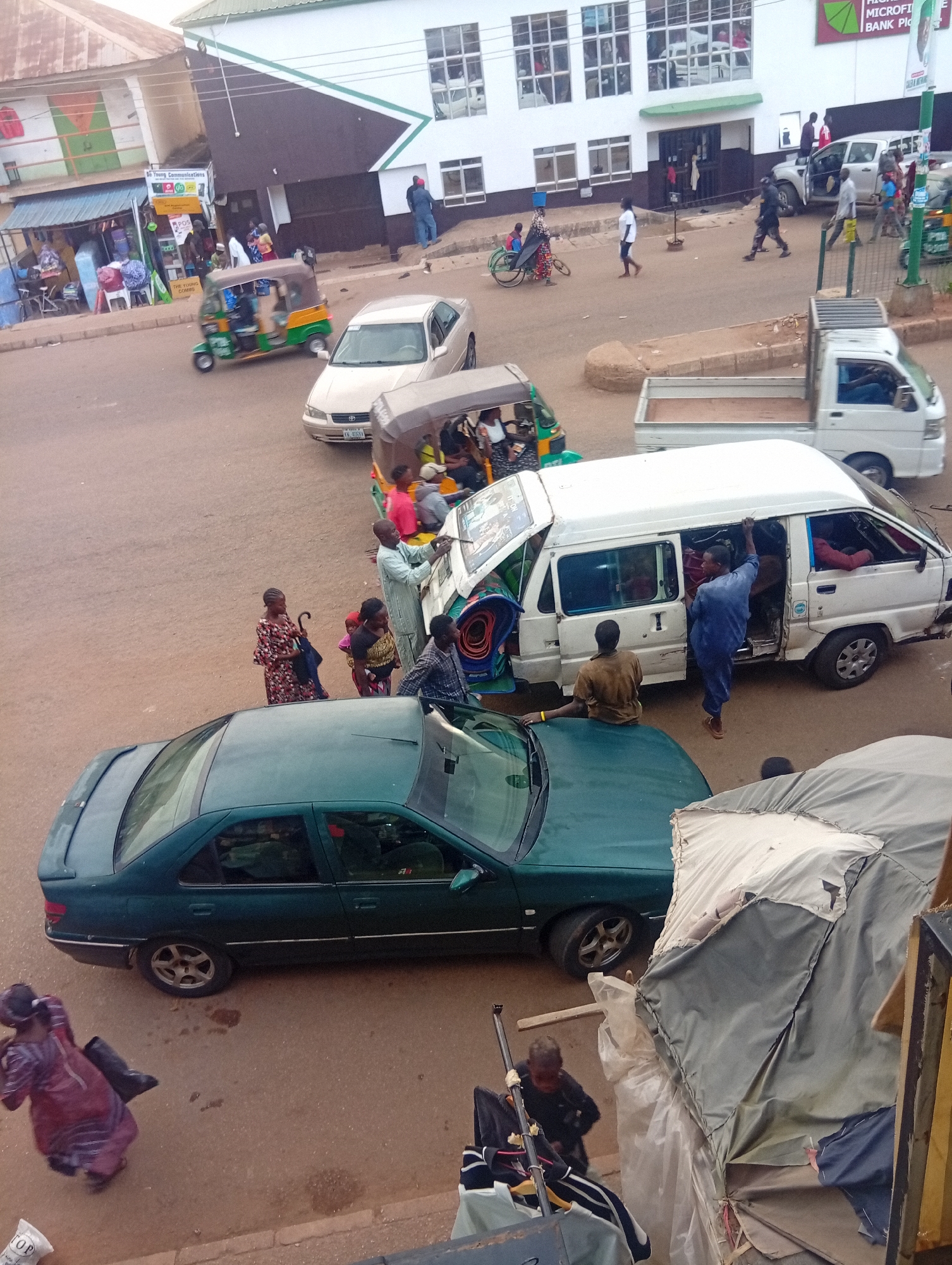 The image depicts a busy street scene in Bukuru, Nigeria, from an elevated perspective. In the foreground, a dark green sedan is parked next to a structure covered with a tattered tent-like material. A woman walks away from the car, carrying an object. A small child is near the tent structure.
In the middle ground, people are gathered around a white van, some interacting with those inside. One person stands in the open doorway of the van, and another is seated on the van’s roof, assisting passengers into an adjacent auto-rickshaw or “keke”. Nearby, a woman with an umbrella and two children stand observing the scene.
Several vehicles, including another auto-rickshaw, a white pickup truck, and a beige sedan, are visible on the road. In the background, pedestrians walk on the sidewalks near buildings. The architecture suggests a mix of residential and commercial structures. On the right side of the frame, signage partially obscures the upper right corner of the image. Street level shops and stalls are visible along the left side of the image.