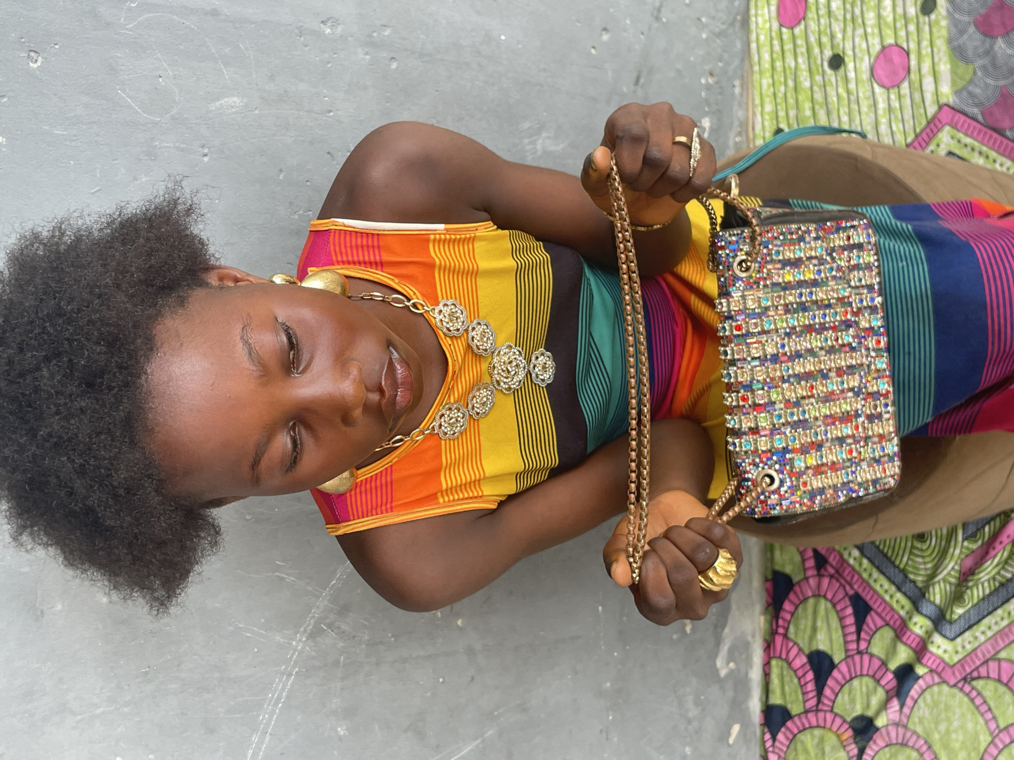 An overhead shot captures a young person with dark skin and natural black curly hair, positioned horizontally across the frame. She wears a vibrant, multi-colored sleeveless top with horizontal stripes in shades of orange, yellow, green, blue, and purple. Her ensemble is accessorized with a prominent gold-toned necklace featuring multiple elaborate, floral-patterned medallions and matching teardrop earrings. Several gold rings adorn her fingers, including a distinctively textured band on her left hand.
The individual is actively engaged, holding a small, rectangular handbag with a gold chain strap. The bag itself is richly embellished with a mosaic of colorful jewels and beads, sparkling under the bright light. She gazes down at the handbag with a slight smile, suggesting contentment or admiration for the item.
The background to the left consists of a plain, light gray, textured wall or ground surface. To the right, a segment of a vibrant, traditional African patterned fabric, featuring green, pink, and black geometric and floral motifs, provides a contrasting splash of color. The bright, even lighting indicates it is daytime. The scene portrays a casual moment in Badagry, Nigeria. There is no visible text in the image.