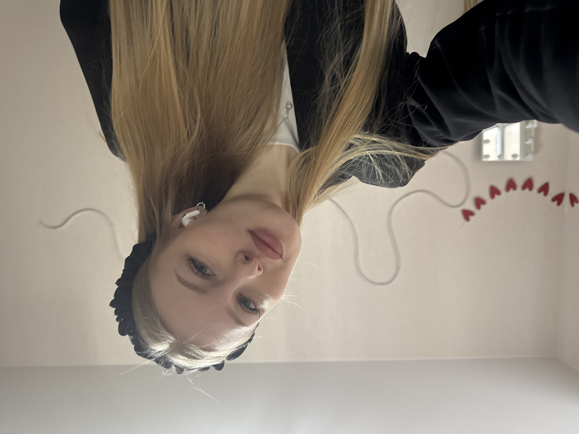 A selfie photograph, taken indoors, depicts a young woman with blonde hair, fair skin, and light-colored eyes. She is wearing a black headband and a black blazer over a white top. A white earbud is visible in her left ear. The background shows a light-colored wall with decorative elements: a string of small red hearts and an electrical cord. A white electrical box is also visible on the wall. The location is not specified in the image, therefore it is impossible to confirm whether this image was taken in Perm, Russia.
