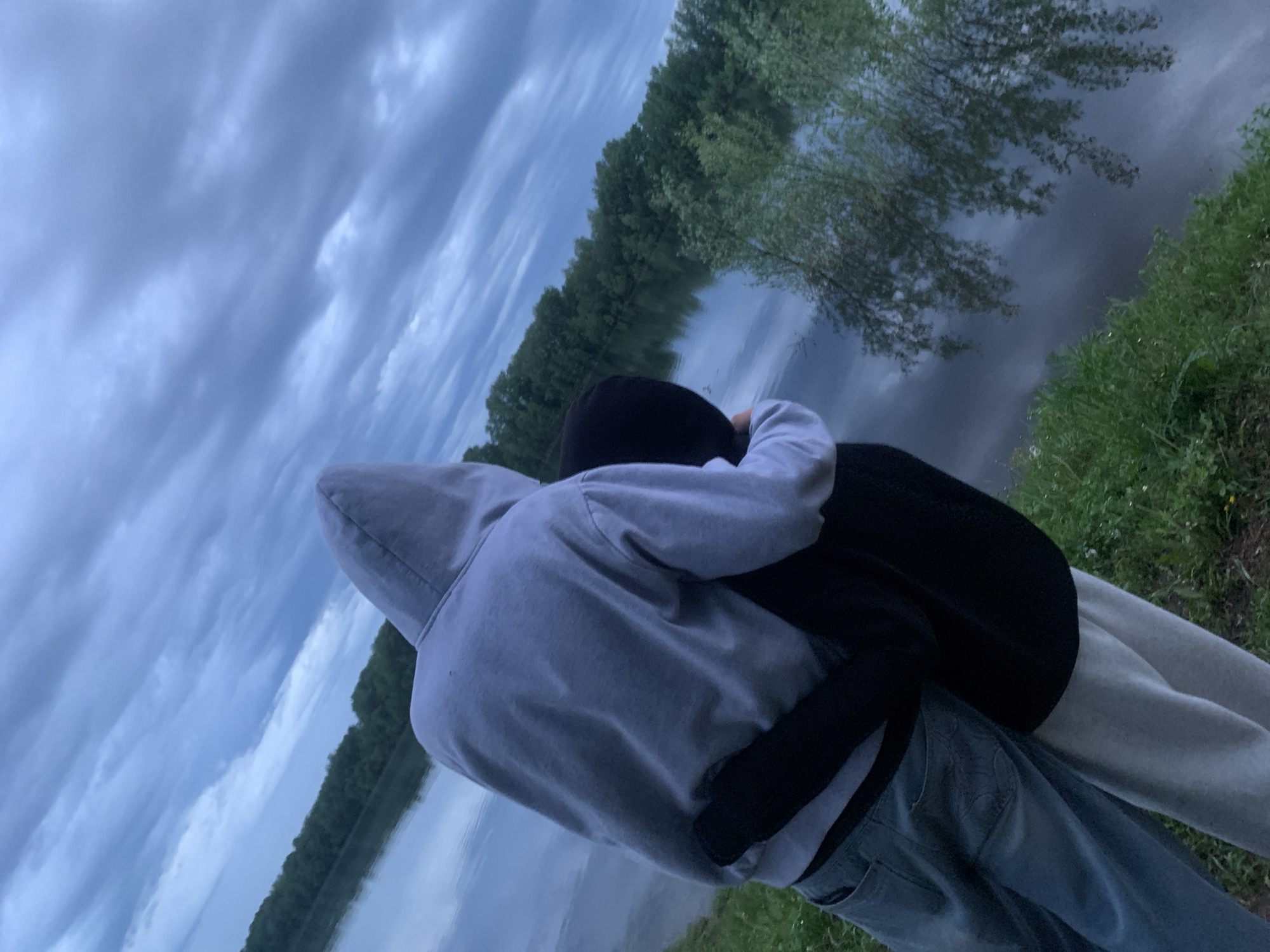 Two individuals stand embraced next to a body of water. The person in the foreground wears a light-colored hooded sweatshirt and blue jeans. The other person wears a dark jacket. The backdrop features a cloudy sky, trees, and the reflection of trees in the water. The scene appears to be outdoors in a natural environment. It's implied that the scene is set at a riverside location near the city of Chaikovsky, Russia.