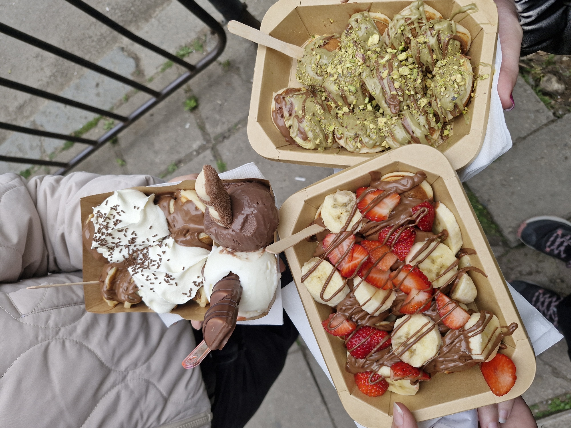 The image provides an overhead perspective, capturing three distinct dessert servings held by individuals outdoors in what is known to be Sofia, Bulgaria. The main subjects are the three brown, disposable paper trays filled with various sweet treats.
In the upper portion, a pair of hands holds a tray brimming with mini pancakes (likely poffertjes) generously slathered with a vibrant green sauce, possibly pistachio-flavored, intertwined with chocolate drizzles and a scattering of crushed pistachios. A small wooden spoon is tucked into this dessert.
To the bottom left, a person wearing a light beige quilted jacket holds a rectangular tray containing waffles or possibly more mini pancakes. This dessert is topped with two scoops of ice cream—one chocolate and one vanilla—alongside whipped cream adorned with chocolate sprinkles and a chocolate-covered confection (resembling a Kinder Bueno). Rich chocolate sauce is drizzled over everything, and a small pink plastic spoon is visible.
The third tray, positioned in the bottom right and held by a hand with manicured nails, showcases mini pancakes abundantly covered with fresh sliced bananas and strawberries, all enveloped in a thick drizzle of chocolate syrup. Another wooden spoon rests within this dessert.
The background reveals a grey, somewhat uneven paved surface, possibly stone or concrete, and the dark horizontal bars of a metal railing. The casual attire of the visible individuals, including a black and white striped sleeve and sneakers in the background, indicates a relaxed, daytime outdoor setting, where friends or family are enjoying these elaborate sweet treats, characteristic of street food or cafe offerings. No visible text is legible on the food containers.