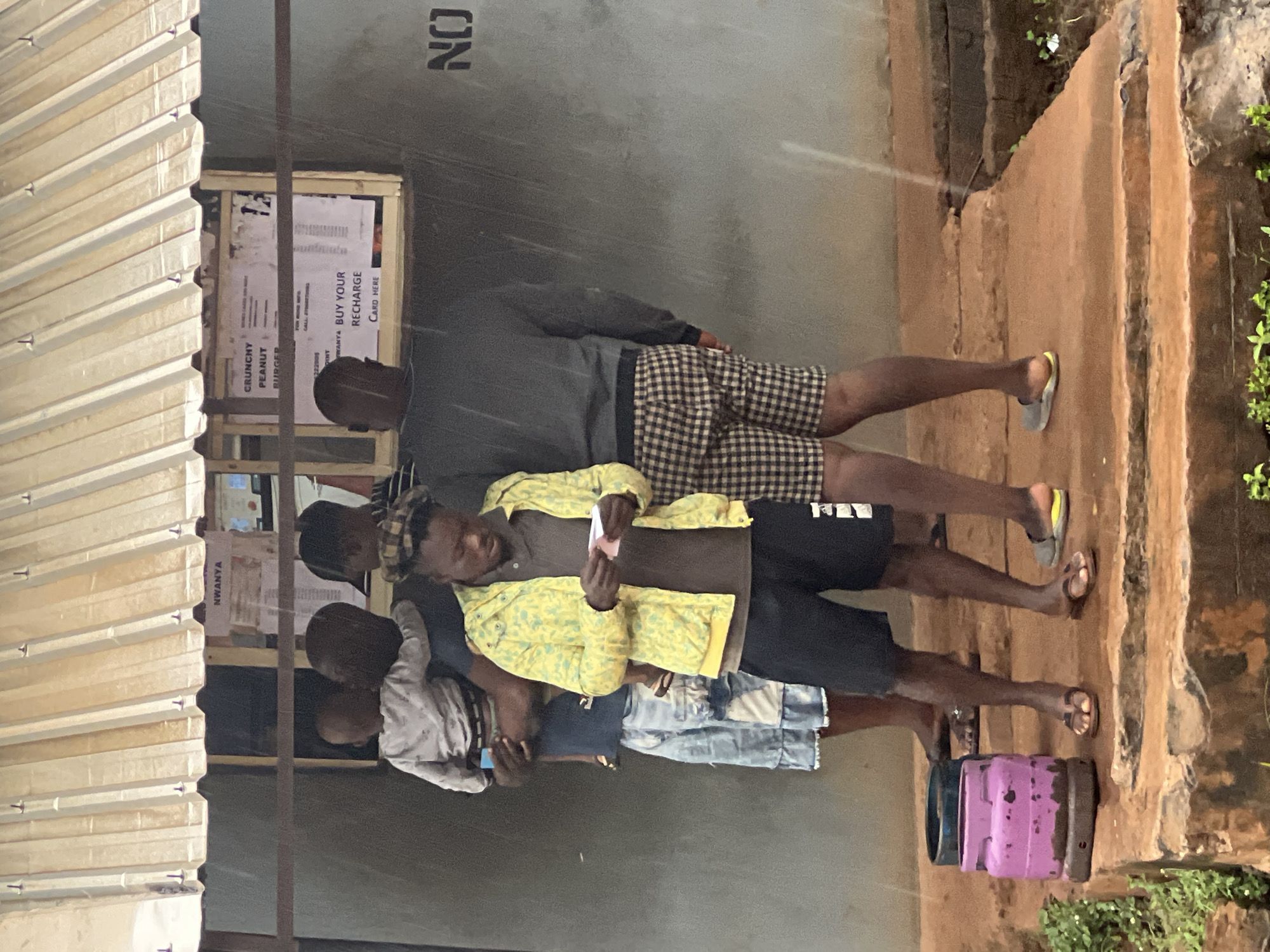 The image depicts a group of young men standing in front of a small shop or kiosk in Nkpor, Nigeria. The setting appears to be outdoors, with a corrugated metal awning overhead and a rough, reddish-brown dirt or concrete pathway with steps in the foreground. The weather seems damp, possibly rainy, as indicated by the wet surfaces and a faint spray of water visible in the air.
The subjects are several young men, mostly dressed in casual clothing like t-shirts, shorts, and a jacket. One individual in the center wears a yellow jacket with a green pattern over a dark shirt and black shorts. Another man to his left is in a light-colored shirt and denim shorts, holding a mobile phone. The man to the right of the central figure wears a dark long-sleeved shirt and checkered shorts, with one foot in a flip-flop. The group appears to be queuing or waiting for something.
Behind the men, a small counter or display area is visible, with some printed materials. One sign, partly obscured, advertises "CRUNCHY PEANUT BURGER" and another reads "BUY YOUR RECHARGE CARD HERE". There is also a large black stencil marking "NO" on a grey wall behind them.
In the foreground, to the right, a small, bright pink propane cylinder is visible, suggesting the location might be a place where gas is sold or used. The overall mood is unremarkable, with no obvious displays of strong emotion. The lighting suggests it could be daytime, but the overcast conditions and possible rain make it difficult to determine the exact time.