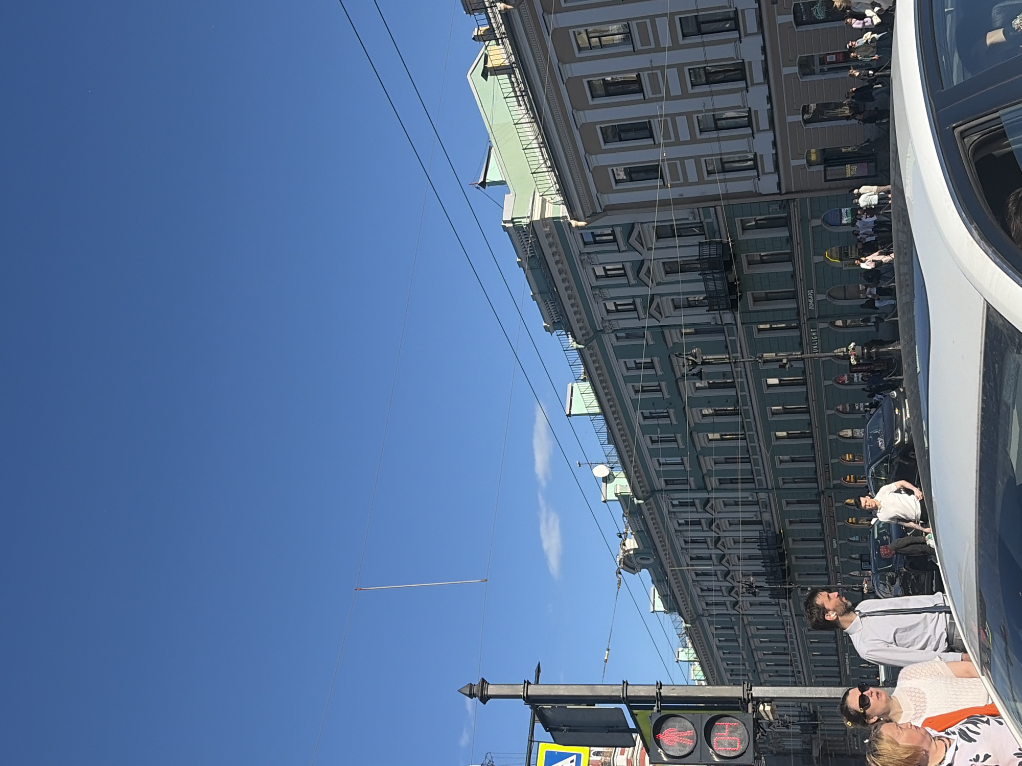 The image captures a street scene in Saint Petersburg, Russia, with a clear blue sky dominating the frame. The perspective is tilted, looking upwards and diagonally along a row of historic buildings lining a street. These buildings feature classic architectural details with multiple stories, intricate window frames, and decorative elements. A bright green facade is visible on one of the buildings, contrasting with lighter colored structures.
Several people are present, standing or walking along the sidewalk in front of the buildings. They appear to be a mix of tourists and locals, engaged in everyday activities. The presence of a traffic light with a red pedestrian signal suggests a street crossing is nearby. Some individuals are dressed in casual summer attire, indicating a pleasant time of day, likely daytime. The overall atmosphere seems calm and ordinary, with no specific event clearly depicted, but rather a snapshot of urban life.
Visible text includes what appears to be a store sign with the word "SUNLIGHT" and possibly "ЛОМБАРД" (LOMBARD), suggesting commercial establishments are located in the buildings. The image conveys a sense of urban exploration and the architectural charm of Saint Petersburg.