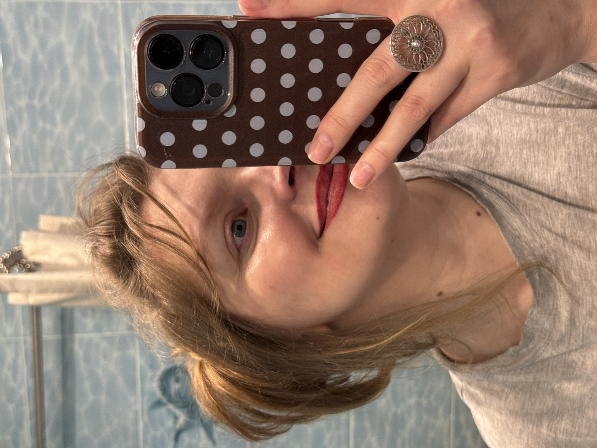 The image is a mirror selfie featuring a young adult female in an indoor setting, likely a bathroom, given the light blue tiled wall in the background. The photo is rotated 90 degrees counter-clockwise.
The subject has light brown hair and blue eyes, and is wearing red lipstick. She is seen playfully sticking out her tongue. She is dressed in a light grey t-shirt. Her left hand holds a smartphone, which has a dark brown case with white polka dots. The phone's multi-lens camera array is visible. On her left ring finger, she wears a decorative silver ring with an intricate floral or mandala pattern.
Behind the subject's reflection, the tiled wall appears to have a subtle pattern. To the left, a white fixture or shelf with some metallic items is partially visible. The lighting is diffused, suggesting it's daytime. The overall activity is a casual mirror selfie, with a lighthearted or playful expression. The location is contextualized as Izhevsk, Russia. No visible text is present in the image.