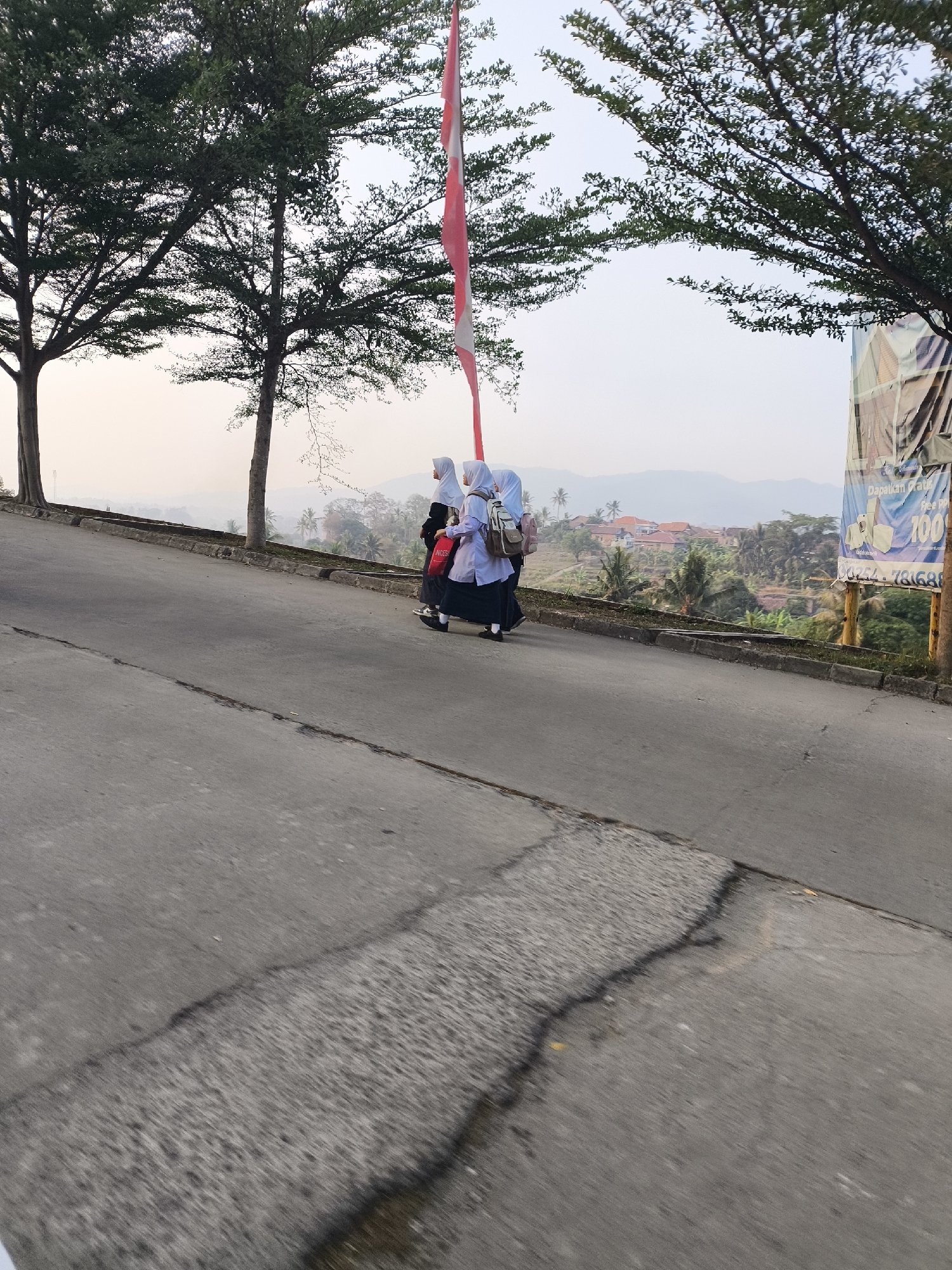 Three young individuals, likely students wearing hijabs and school uniforms, walk uphill on a paved road in Cilegon, Indonesia. The person in the middle carries a light-colored backpack, and the individual behind them has a red bag with the word "NUGS" visible. The road is cracked and appears weathered. Trees with dense green foliage line the left side of the road, and a tall flagpole with a red and white flag is prominently featured. In the background, a hillside community with red-tiled roofs and lush greenery is visible, with hazy mountains in the distance. A large billboard displaying text and images is situated on the right side of the road. The sky is overcast with a pale orange hue, suggesting it is either early morning or late afternoon.