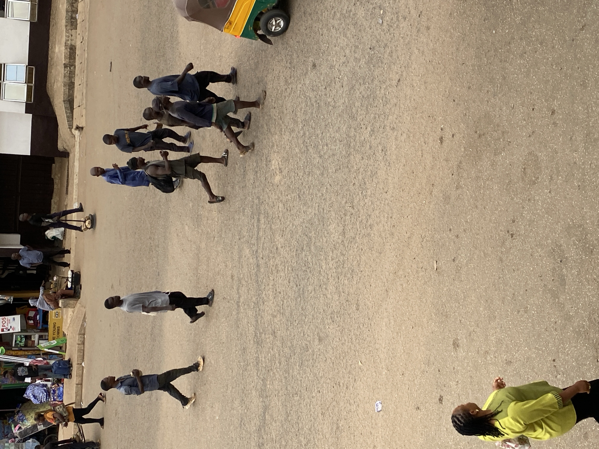 An outdoor scene in Bukuru, Nigeria shows pedestrians and a motorized tricycle on an asphalt street. A group of six men is walking along the road toward the top left of the frame, while other individuals are walking in different directions. One woman in a green shirt is walking in the lower right corner. A row of shops is visible on the left side of the street.