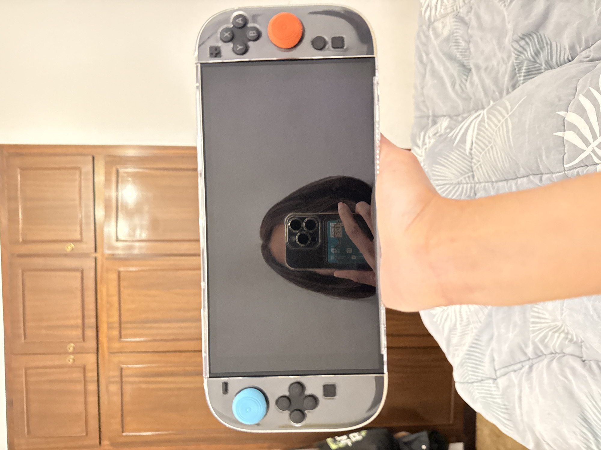A person is holding a Nintendo Switch video game console in front of them. The screen of the console is reflecting the face of the person holding it, including their smartphone. The console has a transparent protective case and blue and orange joystick caps.
Behind the person are:
* A wooden wardrobe or cabinet with multiple panels and brass knobs.
* A bed covered with a gray and white patterned duvet.
* A black bag or other dark-colored objects on the floor.
There is no information in the image that indicates the location is Barcelona, Spain.