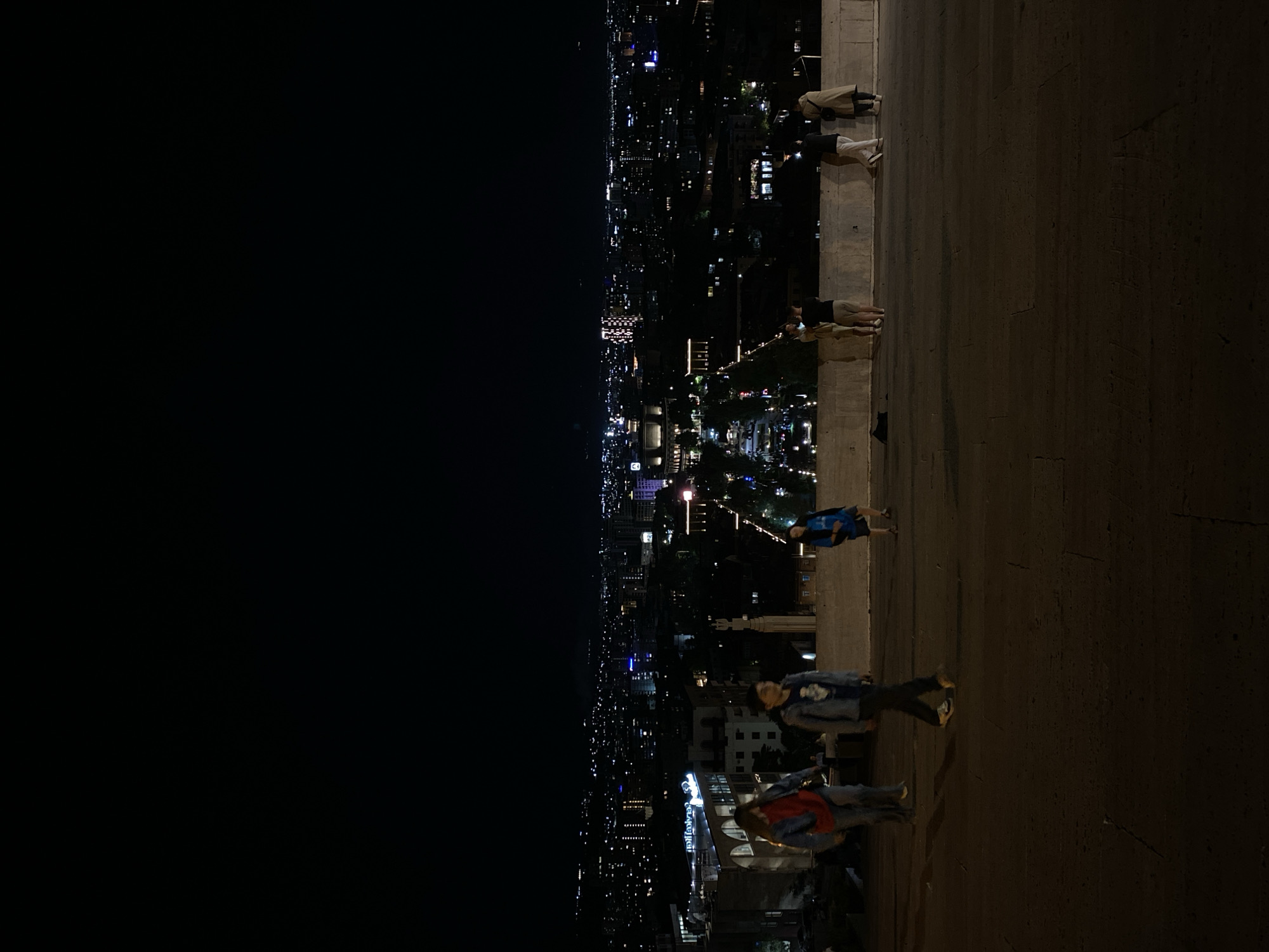 The image displays a nighttime panoramic view of a city from an elevated vantage point. The foreground consists of a dark, textured paved surface, likely an observation deck or pedestrian area, bordered by a low concrete or stone wall.
On this paved area, six individuals are visible. On the left side, two people are present; one wears a red and blue jacket, and the other is dressed in a blue denim jacket with a white design. Towards the right, a person in a blue shirt and dark shorts stands near the wall. Further back on the right, another individual in a light top and dark shorts is positioned by the wall, while two additional people, one in a long light-colored coat and another in a white shirt and light pants, also stand near the wall. The people appear to be observing the city lights, walking, or socializing.
The background is a vast urban landscape illuminated by countless artificial lights under a completely dark sky. Various buildings and streets glow with white, yellow, and blue lights, stretching across the horizon. Several taller structures are distinctly lit, contributing to the cityscape. On one prominent building with a curved facade, illuminated text resembling a hotel name, possibly "Grand Hotel," can be discerned. The time of day is clearly night, with no discernible weather conditions other than clear visibility. The scene conveys a serene atmosphere, with individuals enjoying the expansive nocturnal view of what is identified as Yerevan, Armenia.