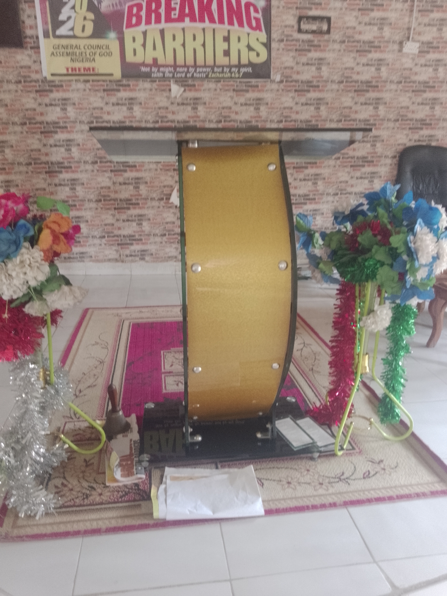 The image shows a podium with a golden, curved body and a clear glass top. The podium is decorated with artificial flowers and tinsel garlands in red, green, and silver. A rug with a floral pattern is placed beneath the podium. On the rug, there are a few papers and a small bell. In the background, there is a brick-patterned wall with a poster that reads "BREAKING BARRIERS" and mentions "GENERAL COUNCIL ASSEMBLIES OF GOD NIGERIA". To the right of the podium, a dark leather chair is partially visible. The scene appears to be indoors, possibly in a church or meeting hall. There are no people visible in the image.