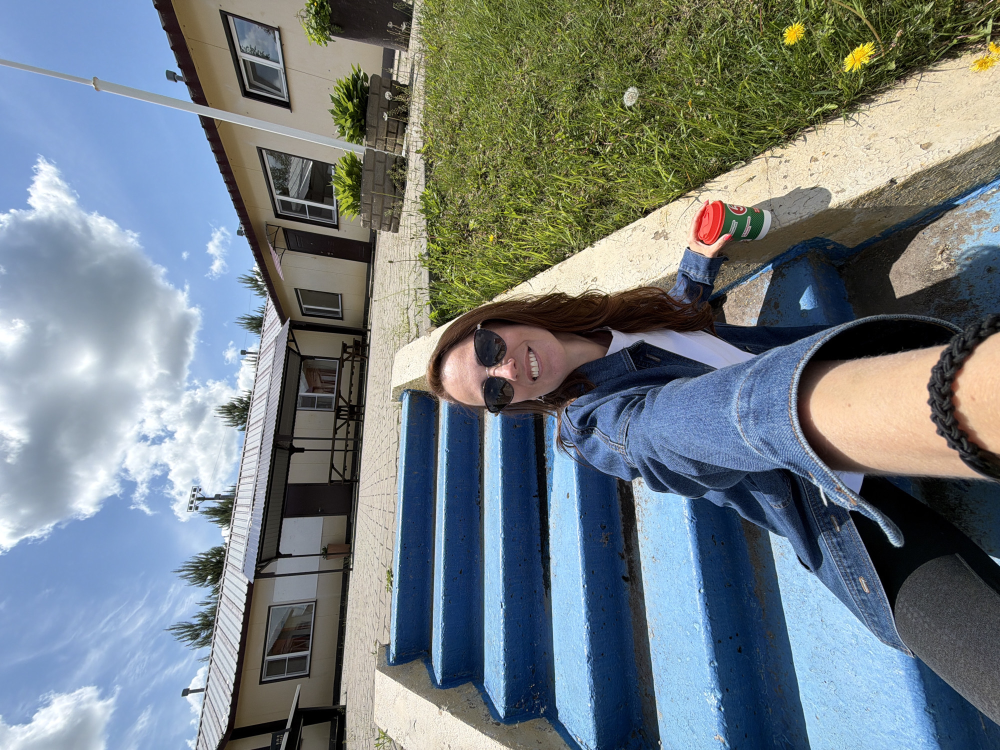 A woman with long, reddish-brown hair and sunglasses is sitting on bright blue stairs outdoors, holding a red and green disposable coffee cup. She is wearing a denim jacket over a white t-shirt and black leggings. Her right arm is extended, suggesting she is taking a selfie. The background features a light-colored building with a metal roof and several windows, surrounded by green grass and some trees. Yellow dandelions are visible in the grass. The sky is blue with white, fluffy clouds. The scene appears to be in a casual, outdoor setting, potentially a park or recreational area, given the presence of picnic tables visible in the cropped images. The style of the building suggests it could be a simple administrative or recreational facility. The location is identified as Birobidzhan, Russia.