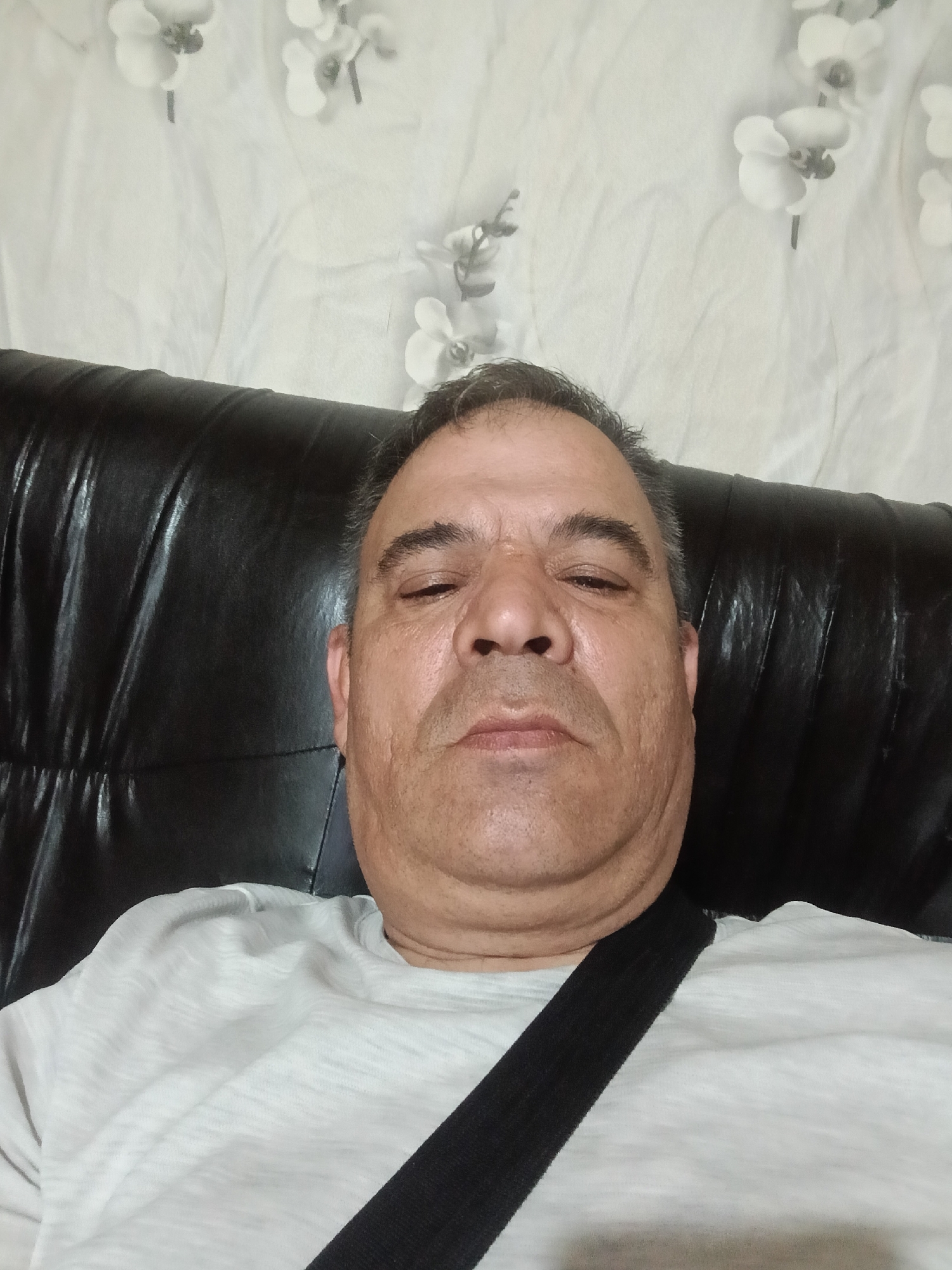 This close-up, indoor photograph features a middle-aged or older man, likely taken in Vetovo, Bulgaria. The man is the primary subject, seated and looking directly at the camera with his eyes partially closed, conveying a relaxed, calm, or possibly tired expression. He has short, dark hair with visible graying, a prominent nose, and a neutral mouth.
He is wearing a light-colored, possibly white or light gray, subtly textured t-shirt. A black strap, resembling a seatbelt or a bag strap, runs diagonally across his chest from his right shoulder down to his left side.
The man is positioned in front of a dark, likely black leather, piece of furniture, such as a sofa or chair, which appears textured and somewhat creased. Behind the furniture, the background consists of a light-colored, possibly white or off-white, wall or fabric with a delicate pattern. This pattern features stylized white floral motifs, resembling orchids, with dark centers and stems, sparsely distributed across the light surface. The lighting in the image suggests an indoor setting, with no indication of the specific time of day or outdoor weather conditions. There is no visible text in the image.