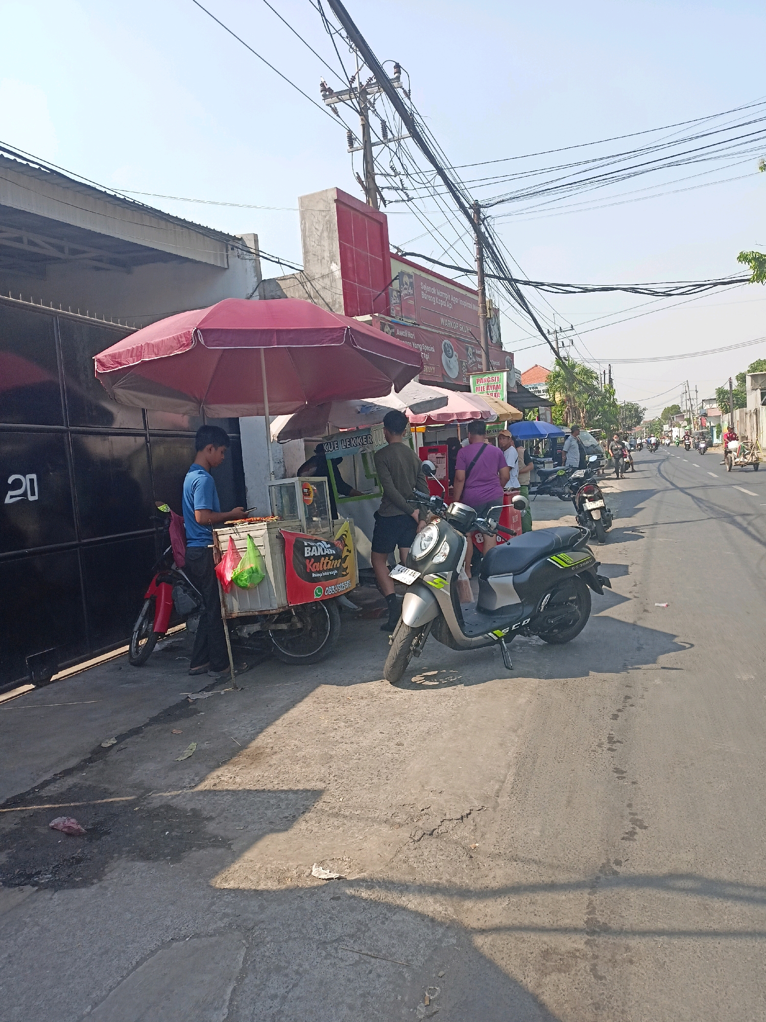 The image depicts a street scene in Surabaya, Indonesia, under a clear, bright sky. The foreground is dominated by a street food stall selling what appears to be grilled food. A young male in a blue shirt is attending to the stall, which is equipped with a display case and a cart on wheels. A red motorcycle is parked next to the stall. Several other motorbikes and a rickshaw are visible on the street, suggesting a typical urban environment with traffic. The background features a building with a dark metal gate marked with the number "20" on the left. Further down the street, more shops and stalls are lined up, some with awnings and signs. Power lines are prominently strung overhead. The ground is paved, with some areas of unevenness and a few pieces of debris. The overall impression is one of a bustling daytime street market.