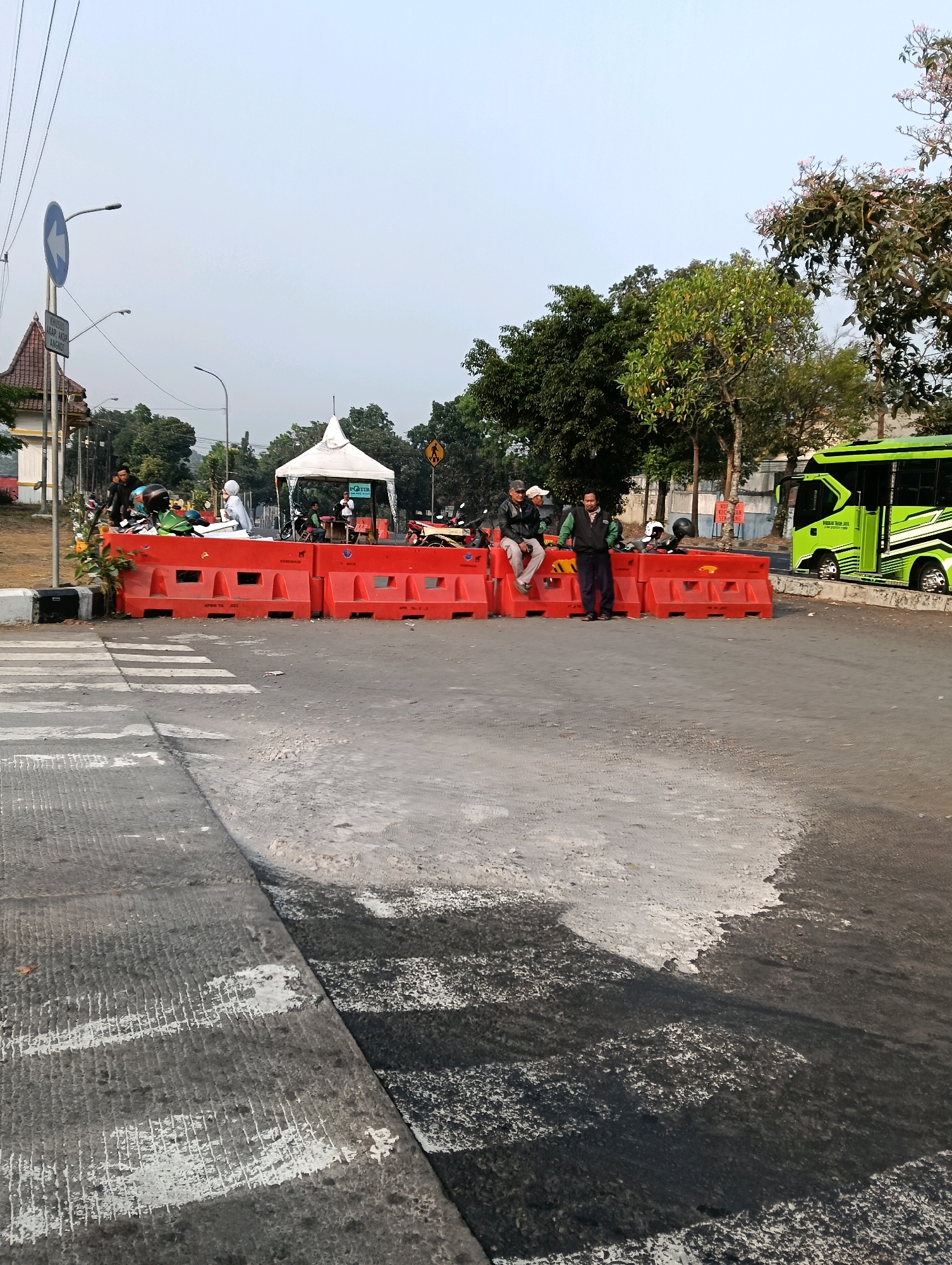 The image depicts a street scene in Magelang, Indonesia, characterized by a temporary blockade of red plastic barriers spanning across the road. Several motorcycles are parked behind the barriers, and a white tent structure is visible. Three men are positioned near the barriers; one is seated, another is standing, and the third is leaning against a motorcycle. A green and black bus is parked on the right side of the frame. The foreground shows a pedestrian crossing with worn-out markings. The background includes trees and utility poles, suggesting an urban or semi-urban environment. The sky is overcast.