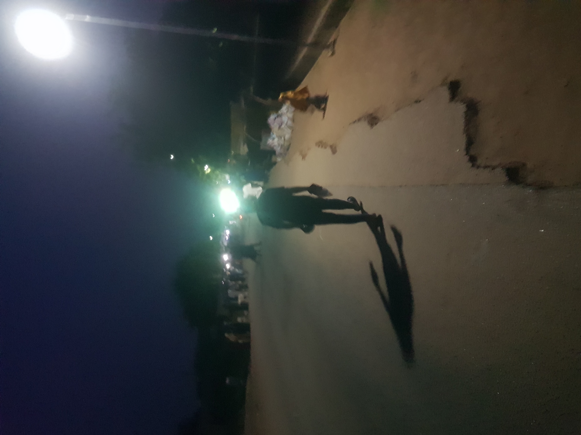 A dimly lit street scene at night in Dabawa, Nigeria, features a pedestrian walking under a street light. The pedestrian, silhouetted by the light, casts a long shadow on the pavement. Other streetlights and blurred figures are visible in the background, suggesting a populated area. The road surface is uneven. Rubbish is piled up to the side. The sky is dark.