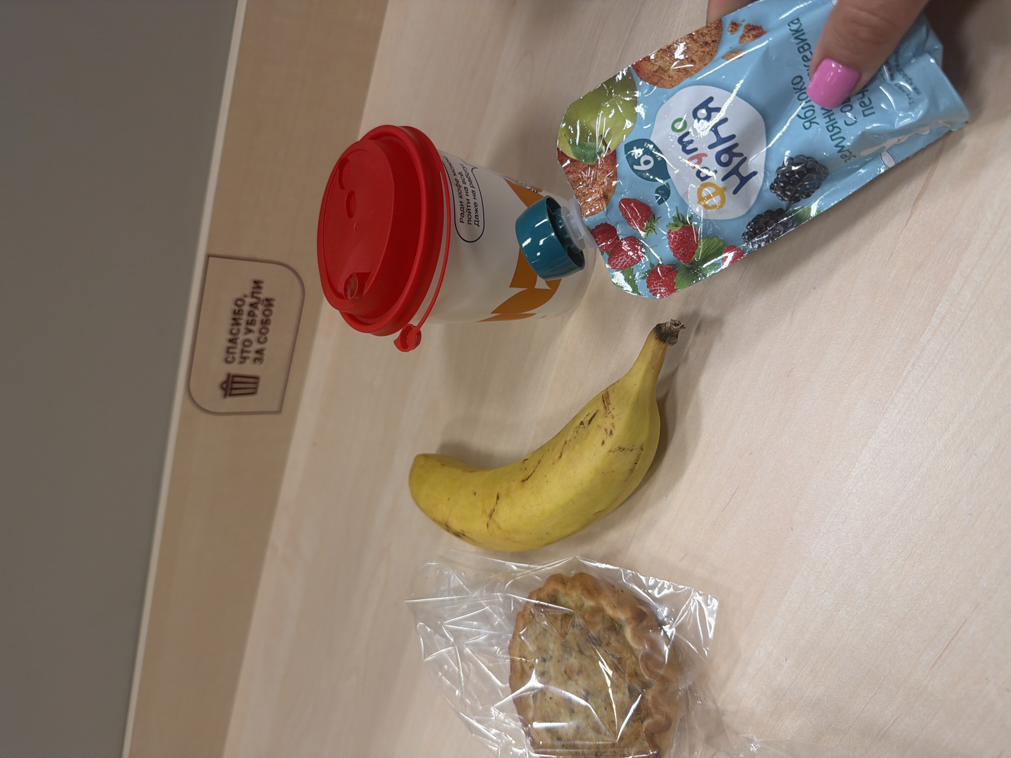 This image shows a table with several items on it: a reusable coffee cup with a red lid, a baby food pouch, a banana, and a wrapped pastry. The baby food pouch is being held by a hand with pink fingernails. There is also a trash can icon and text in Russian on the table's surface. The table appears to be made of light-colored wood or laminate.
