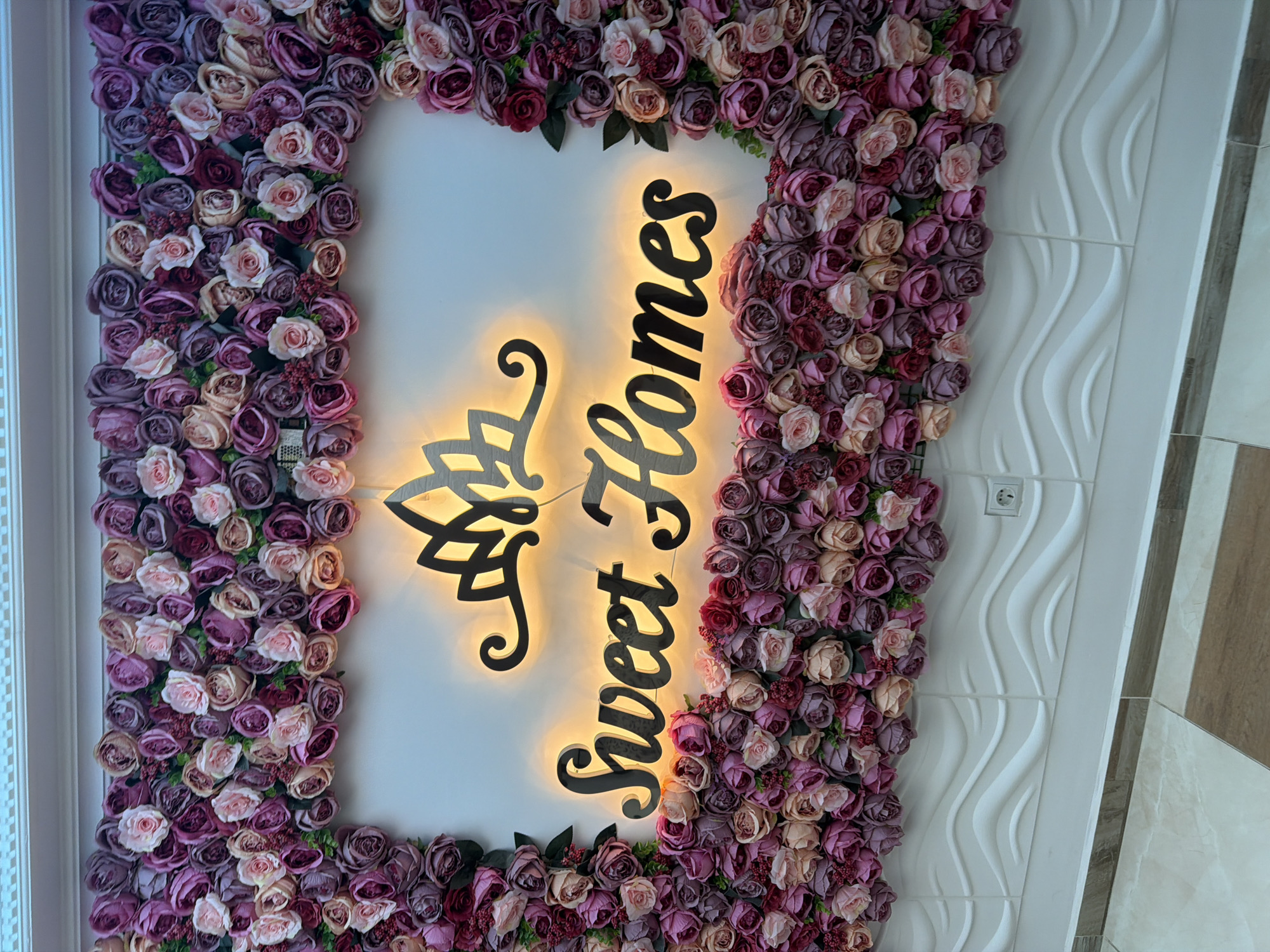 This image shows an illuminated sign that says "Sweet Homes" inside a floral arrangement. The sign is centered and has a crown-like graphic above it. The floral arrangement is made up of various shades of pink and purple roses. The sign and flowers are mounted on a white wall with a subtle wave-like texture at the bottom. The floor below is tiled with a light beige and brown pattern. The image appears to be taken from a slightly upward angle. The location is identified as Nesebar, Bulgaria.