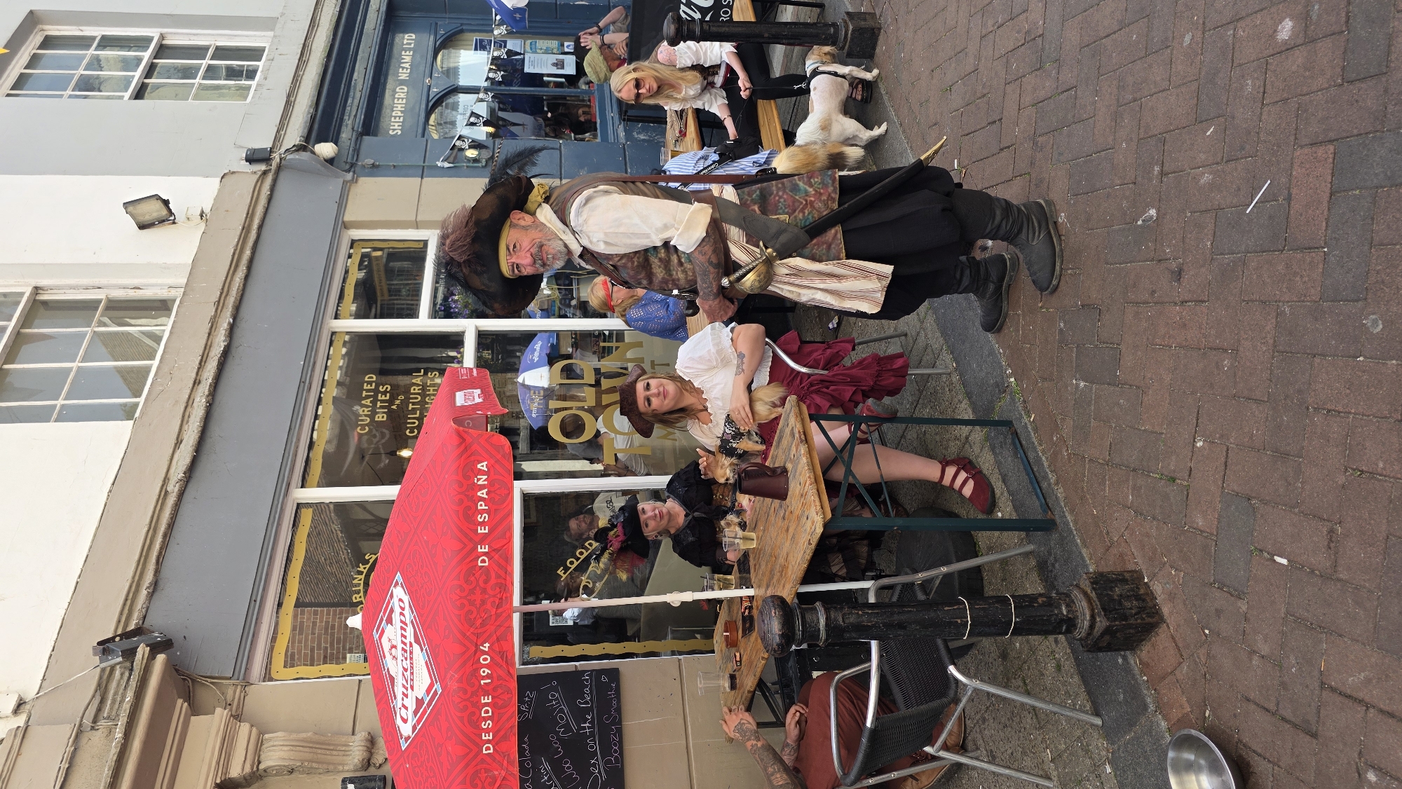 In St Leonards, United Kingdom, a group of people dressed in pirate-themed attire are gathered outside a cafe. A man in a prominent pirate costume, complete with a tricorn hat, ruffled shirt, vest, and sword, stands at the forefront of the image, looking directly at the camera. To his left, seated at a wooden table under a red umbrella, are two women also in pirate-themed outfits. One woman holds a small dog, while the other has a dog resting on her lap. Several drinks are visible on the table. A dog in a harness is standing near the seated women. The cafe appears to be named "Old Town". The setting is an outdoor seating area with brick paving and tables and chairs. The building facade behind the cafe has windows and signage.
