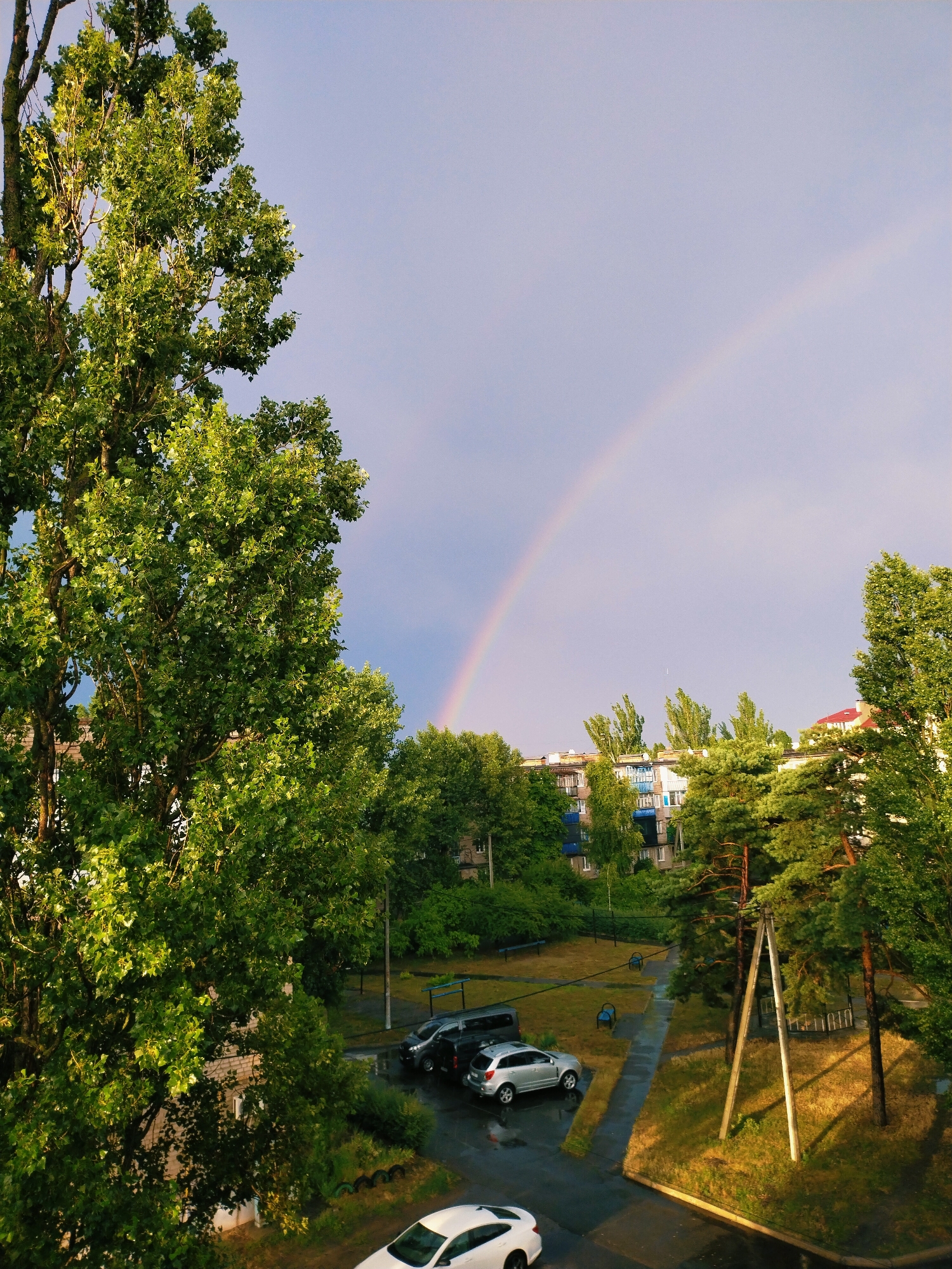 This is an aerial, medium shot of a residential area in Horishni Plavni, Ukraine, after a rain shower. The sky is overcast with a faint rainbow arching across the upper right quadrant of the frame. In the foreground, a large deciduous tree with vibrant green foliage dominates the left side of the image. Adjacent to it, the side of a brick building is visible.
The middle ground reveals a paved courtyard with several parked cars, including a silver SUV, a dark minivan, and another dark car. The pavement appears wet, indicating recent rainfall. Patches of grass and scattered benches are visible in the courtyard, along with a children's playground structure. Pine trees and other greenery are interspersed throughout the scene.
In the background, multi-story apartment buildings with light-colored facades and blue window frames are visible, characteristic of Soviet-era architecture common in Ukraine. The overall atmosphere is serene, with the lingering presence of rain adding a fresh quality to the environment. No people are visible in the frame.