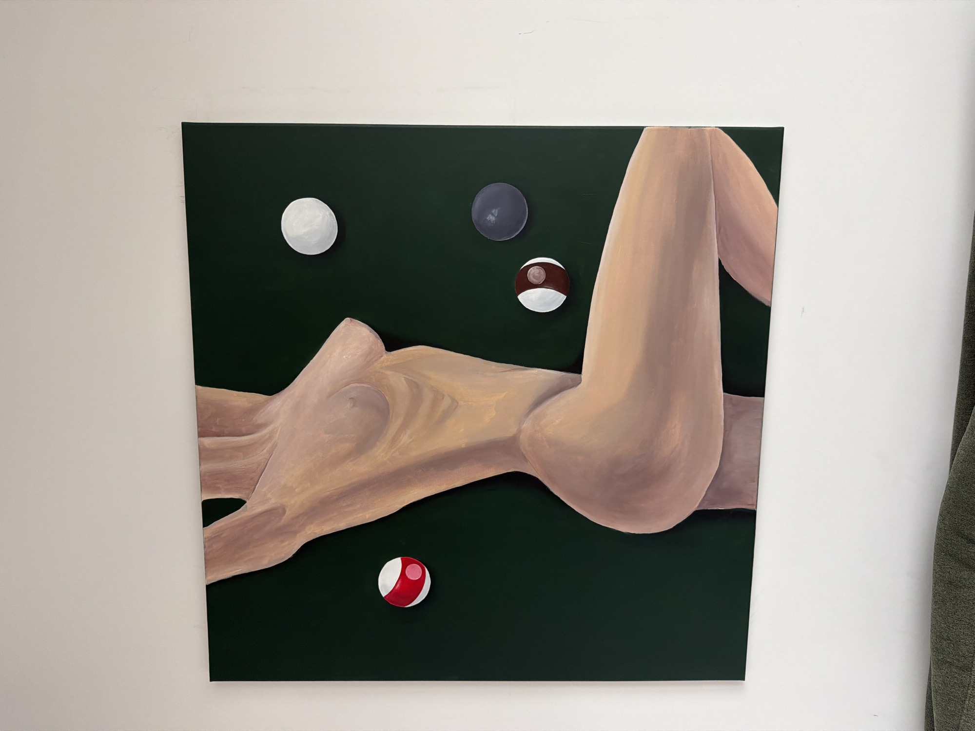 A square painting hangs on a white wall. The painting depicts a reclining nude figure rendered in muted flesh tones against a dark green background. Scattered around the figure are four billiard balls of various colors: a solid white ball, a solid grey ball, a ball with brown stripes, and a ball with red stripes. A partially visible green curtain or fabric hangs on the right side of the image, adjacent to the painting. The location of the image is unknown and cannot be inferred. There is no evidence to suggest this painting is located in Warsaw, Poland.