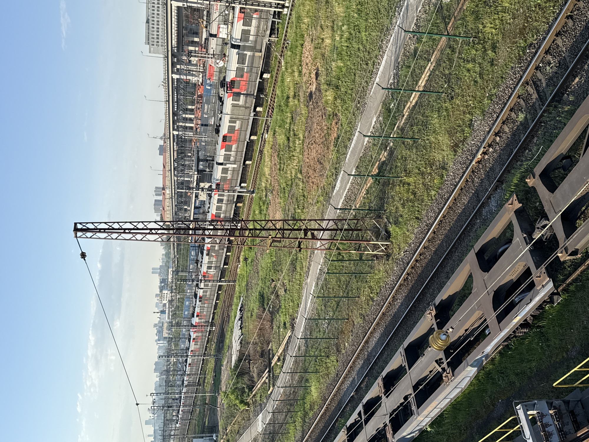The image shows a railway yard in Moscow, Russia. The sky is light blue with some clouds. A cityscape of tall buildings is visible in the distance. Multiple railway tracks run through the scene, with a freight train car visible in the lower right corner. Some commuter trains, painted in grey and red, are parked on the tracks to the left. A metal utility pole spans the tracks with overhead electrical wires. A green barrier fence separates the tracks from the grassy land around the yard.