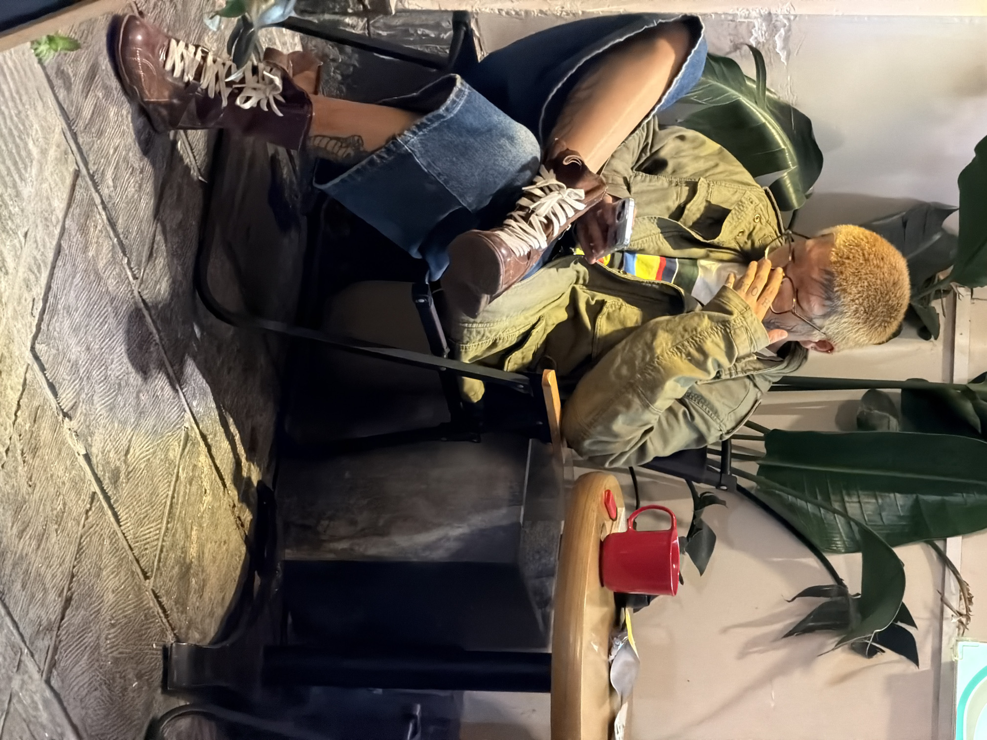 A person with short, bleached blonde hair wearing glasses, a green jacket, and blue jeans is seated on a black chair, holding a smartphone. They are resting their head on their hand, appearing contemplative. To their right, a round wooden table holds a red mug, a spoon, and what appears to be a packet of sugar. In the background, large green plants with broad leaves are visible against a light-colored wall. The person's feet are clad in brown, lace-up boots. The floor is tiled with a wood-like pattern. The scene is indoors, possibly a cafe or a waiting area, given the furniture and plants. There is no explicit indication of the location being in Yuzhong District, China, based solely on the visual information provided.