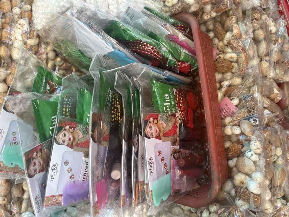 The image shows a collection of various items arranged in a stack in a plastic basket. The items are packaged in clear plastic bags with labels and are likely for sale at a market stall in Panchkula, India. The products include various beauty products, including Patanjali brand items, henna, and a comb, as well as some bracelets or bangles. There are also many small, white, and brown objects visible in the bottom of the image, which are likely nuts or seeds. The items are arranged neatly in a stack and there are no people visible in the image. The image is likely taken from a stall at a market in Panchkula, India.