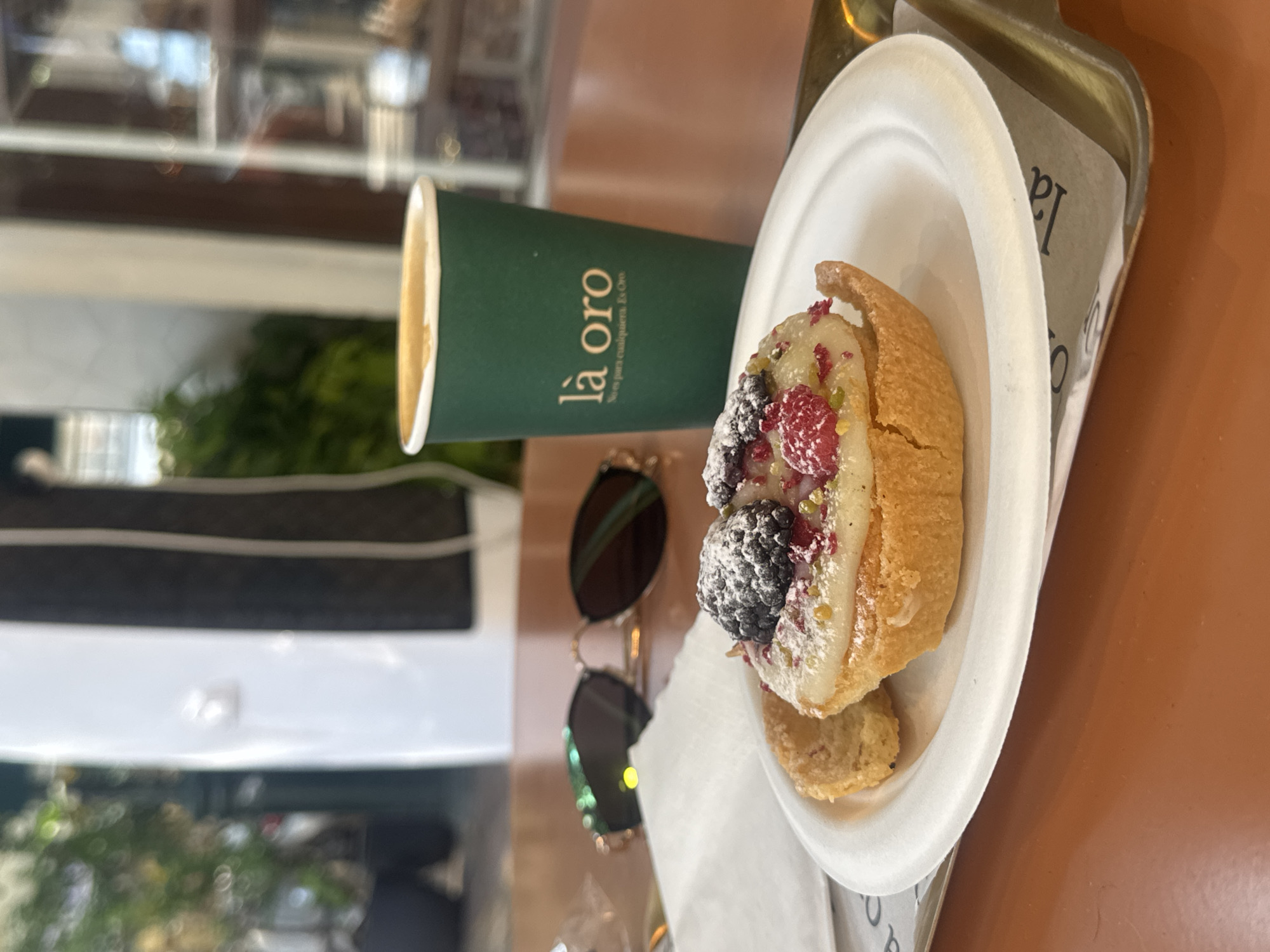 The image depicts a close-up view of a small, open-faced pastry with berries and powdered sugar, placed on a white paper plate. To the left of the pastry, a small cookie is visible. A green paper coffee cup with a white rim and the logo "là oro" is positioned behind the plate, slightly to the right. The coffee cup appears to contain a light-colored beverage. In the background, a pair of dark-lensed sunglasses with a rose-gold frame rests on an orange-brown surface, likely a table. The scene is set outdoors, with a blurred background that suggests a cafe or restaurant setting with greenery and architectural elements, consistent with a location in Ramat Gan, Israel. The overall impression is of a casual outdoor dining setting.