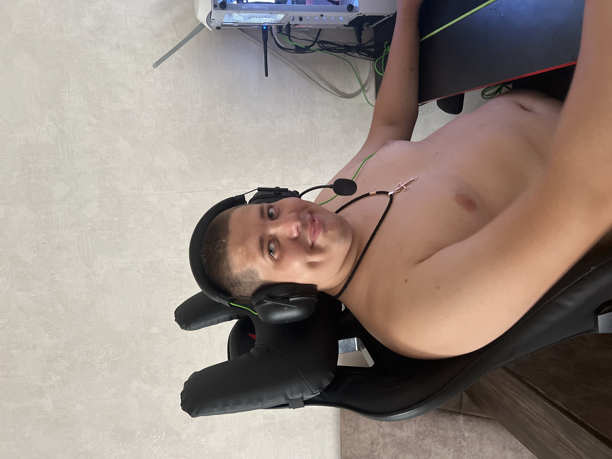 The media file shows a young man sitting in a gaming chair, facing the camera. He is shirtless and wearing headphones with a microphone. He has a short haircut and appears to be in his early twenties. A necklace with a cross pendant is visible around his neck. Behind him, a computer tower and a desk with cables are visible. The background consists of a light-colored, textured wall. The lighting suggests it is daytime, and there are no clear indications of the weather or specific location beyond the provided context of Марковский, Russia. The man has a neutral expression, and the overall scene appears to be indoors in a room set up for gaming or computer use.