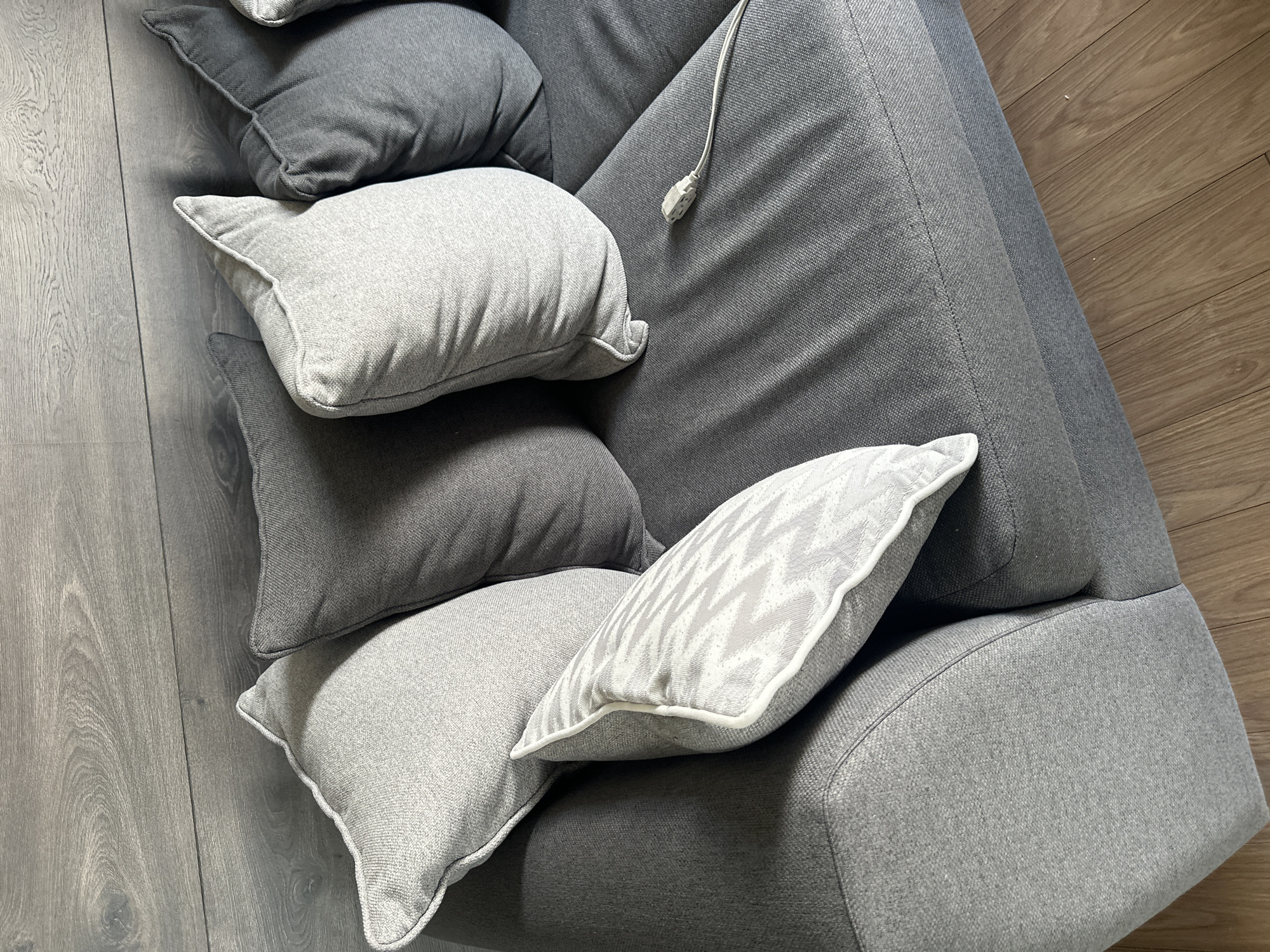 The image shows a corner of a gray sofa with several throw pillows. The pillows are various shades of gray and textures, with one pillow featuring a light gray zigzag pattern with a white trim. A white electrical cord with a plug is visible on the sofa. The flooring is a light brown wood-grain. The scene is interior and has no visible people or actions. There is no discernible information to suggest this image was taken in Huixquilucan de Degollado, Mexico, or any other city.