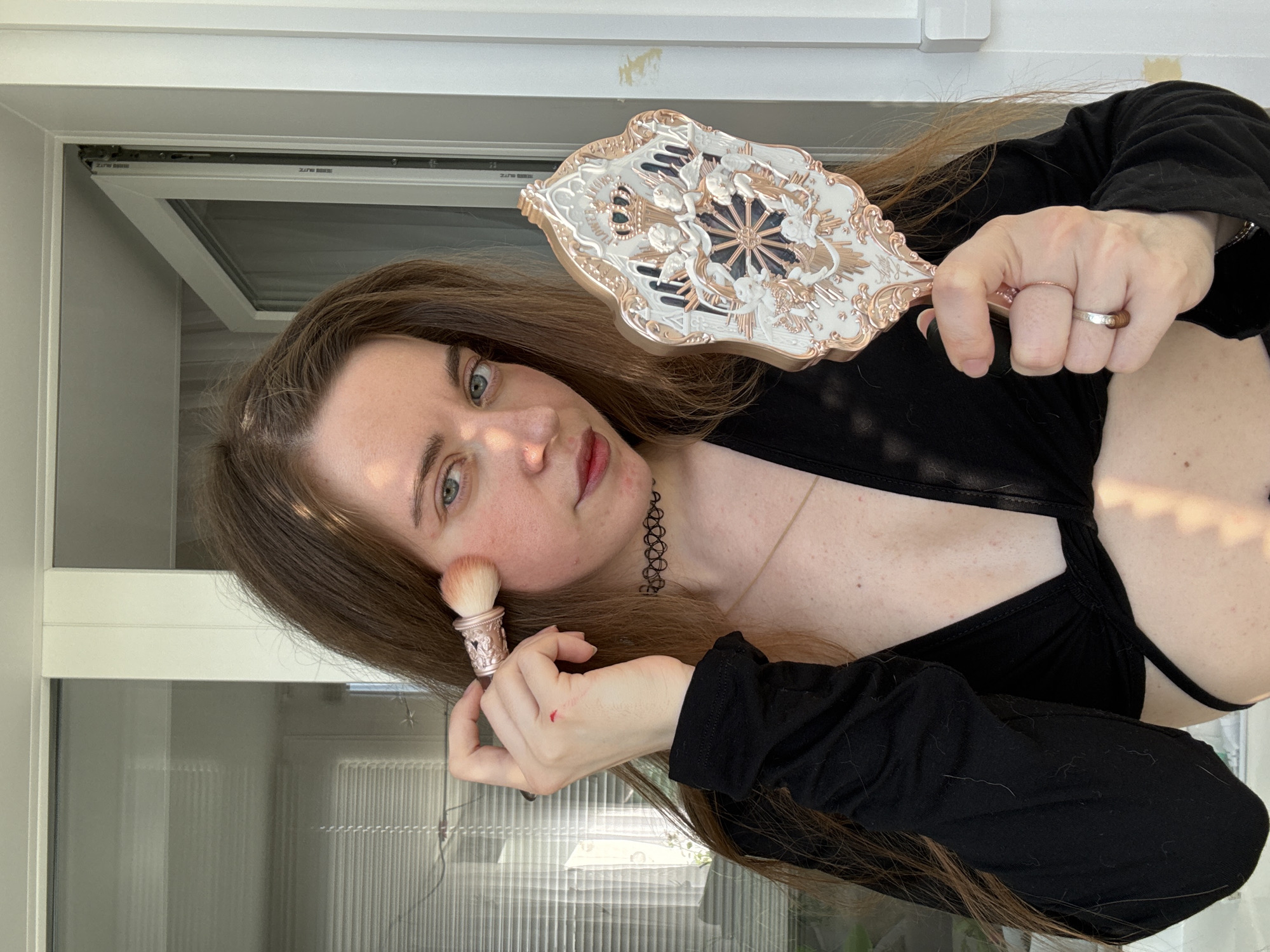 A person with long, brown hair is applying makeup in a selfie. They are wearing a black top and a choker necklace. They are holding a makeup brush to their cheek and a decorative mirror in their other hand. The background appears to be an indoor setting with large windows, suggesting it might be daytime. The person's expression is neutral, and the overall mood is relaxed, as if they are getting ready.