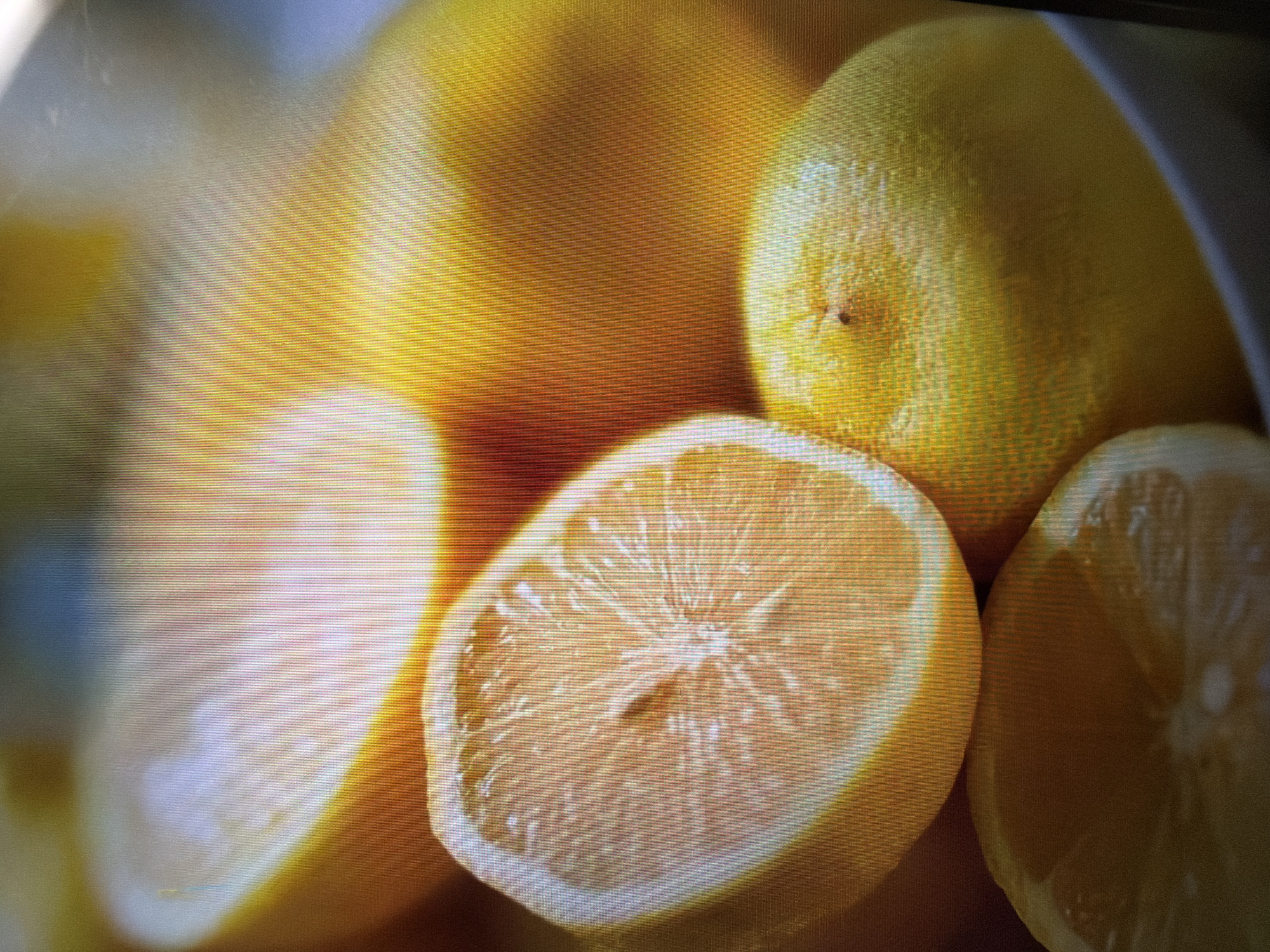 The image is a close-up, slightly out-of-focus shot of several lemons, some whole and one halved. The lemons are bright yellow, with visible texture on their skins. The halved lemon reveals its juicy interior with segments and pith. The lighting creates highlights and shadows, emphasizing the roundness and form of the fruit. The background is a soft blur of yellow and white. The image quality suggests it was taken on a screen, possibly a monitor or television, with visible pixelation and a grid-like pattern across the surface. The context of Moscow, Russia, is not discernible from the visual content of the image itself, as it depicts a simple arrangement of fruit in what appears to be an indoor setting.