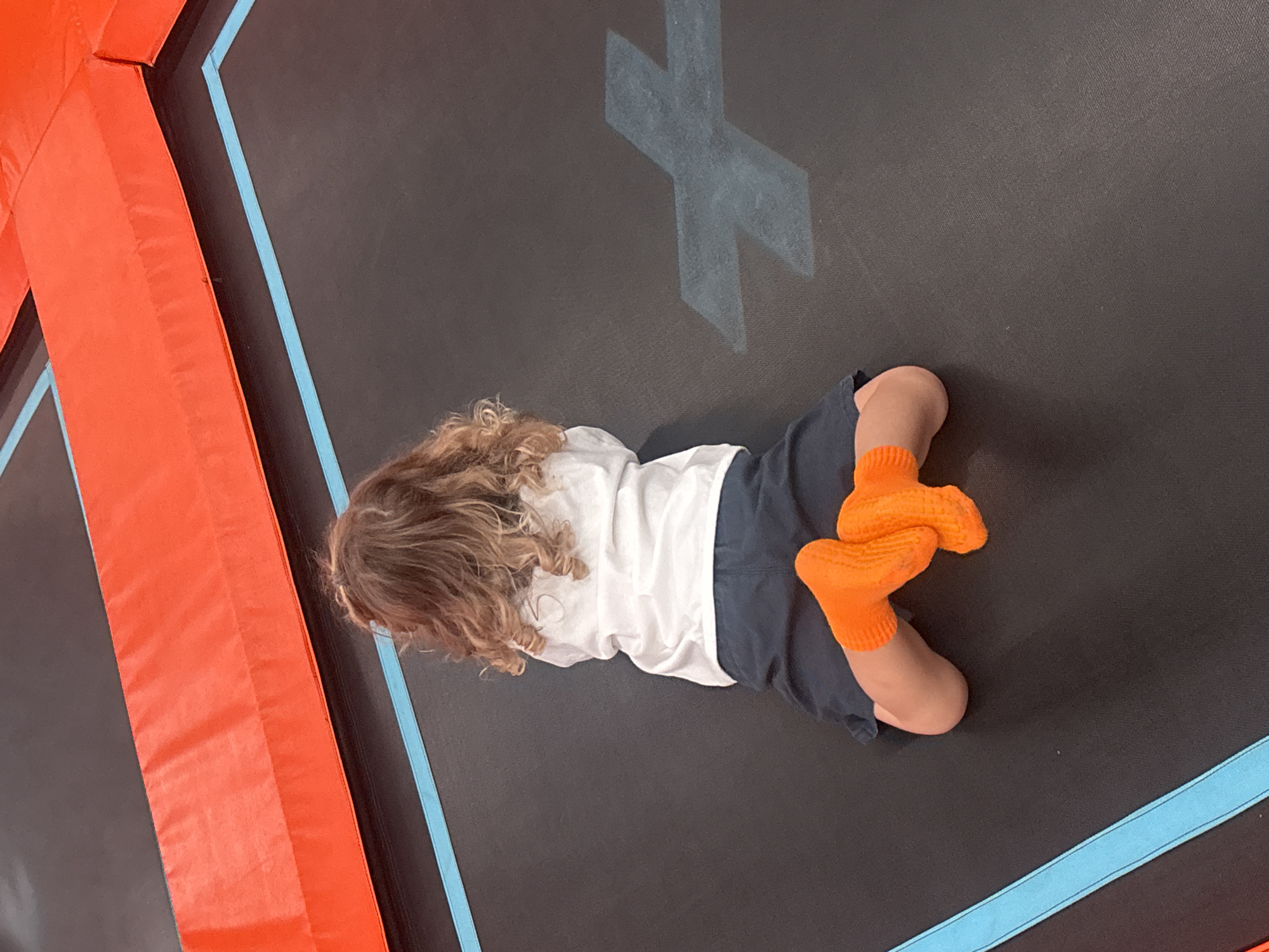 The image shows the back of a young child with curly brown hair lying on a trampoline. The child is wearing a white long-sleeved shirt and navy blue shorts. Their feet are in bright orange socks. The trampoline has a black mat with blue and orange padded edges. A large, light gray "X" graphic is visible on the trampoline mat to the right of the child. The overall scene suggests a recreational indoor trampoline park. There is no specific visual information within the image to determine the exact location within Dubai, United Arab Emirates, other than it is an indoor trampoline park.