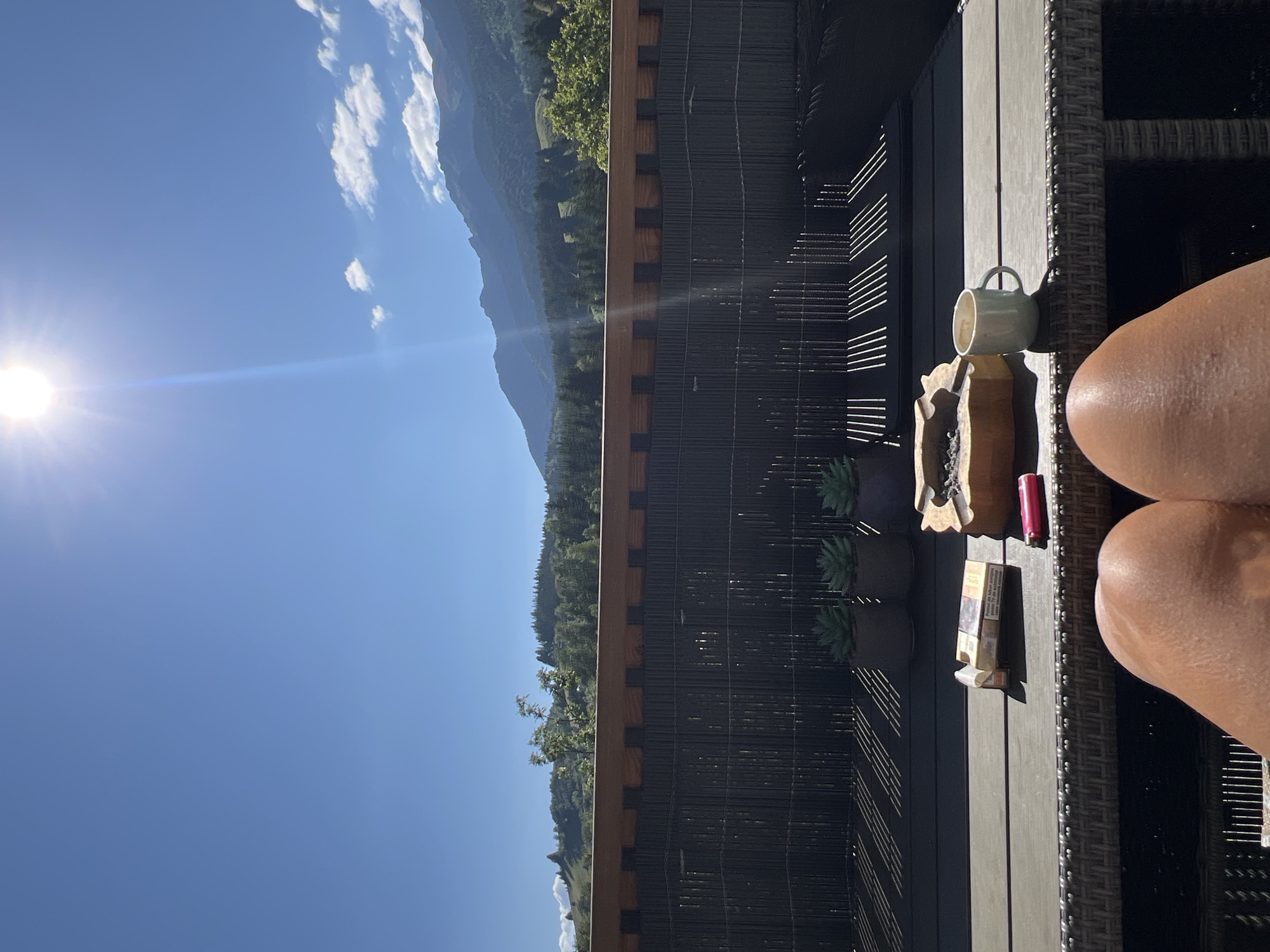 A person's bare legs are visible in the foreground of the image, positioned in front of a table. On the table, there is an open pack of cigarettes, a pink lighter, an ashtray containing ashes, and a white mug. Behind the table, there is a dark-colored fence or partition, and beyond that, a lush green landscape of trees and mountains under a bright, sunny sky. The sun is high in the sky, creating lens flare and casting distinct shadows on the table and ground. The overall scene suggests an outdoor setting, possibly a balcony or patio, enjoying a sunny day.