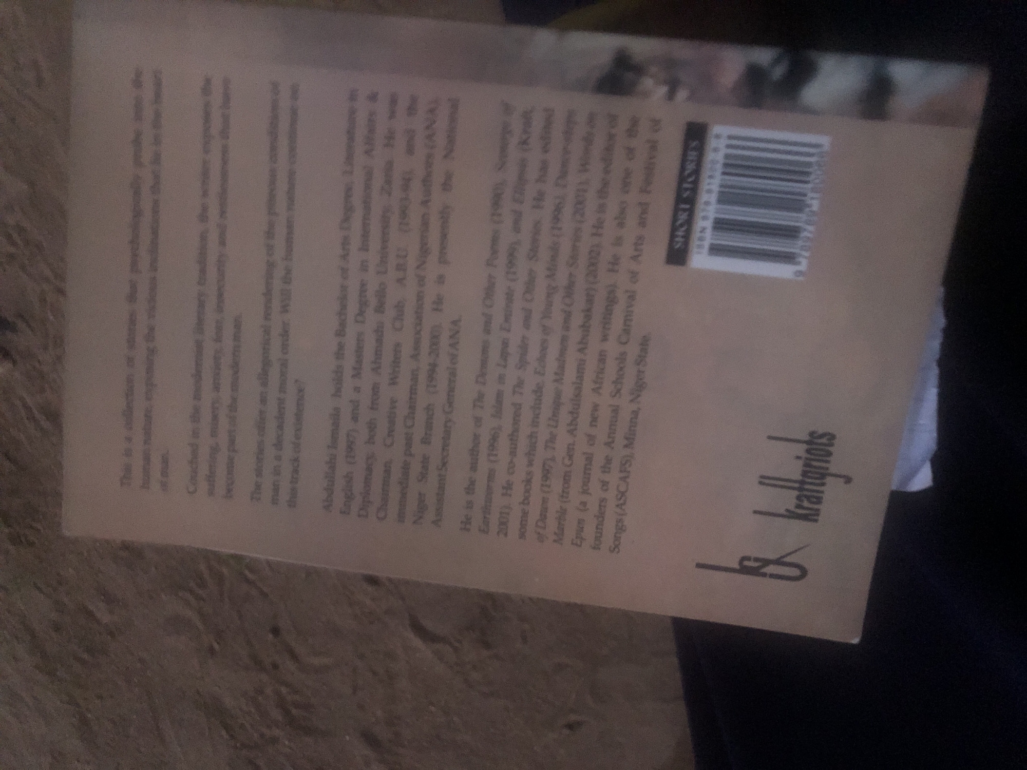 The image shows the back cover of a book, likely a collection of short stories. The text describes the author, Abdullahi Ismaila, and mentions his academic background and previous works. It also lists "SHORT STORIES" above a barcode and an ISBN number. The lower left corner of the book cover features a logo that appears to be "bk kraftgriots". The image is taken from a low angle and the lighting is dim, with the majority of the scene in shadow. There is no discernible information about the city of Jalingo, Nigeria, within the visible text or imagery.