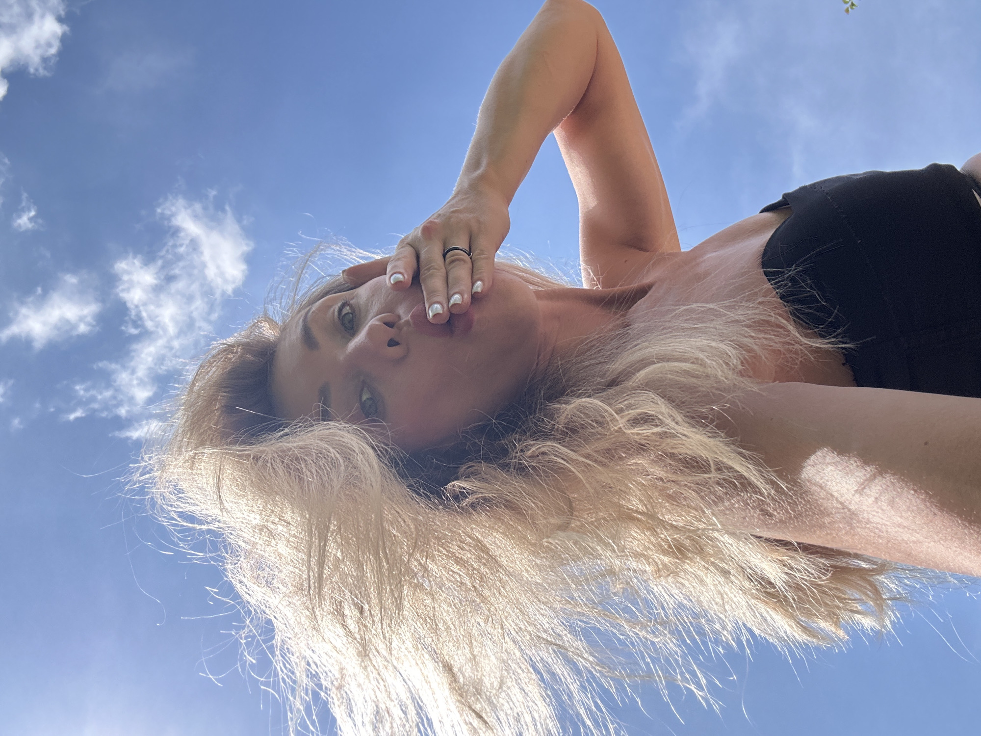 An outdoor, low-angle, close-up selfie captures a blonde woman in the city of Armavir, Russia, as she blows a kiss to the camera. The woman, who is wearing a strapless black top, has her right hand covering her mouth. Her hair is being blown by a strong wind, obscuring a large portion of her face. The background is a bright blue sky with scattered white clouds. The lighting suggests it is daytime.