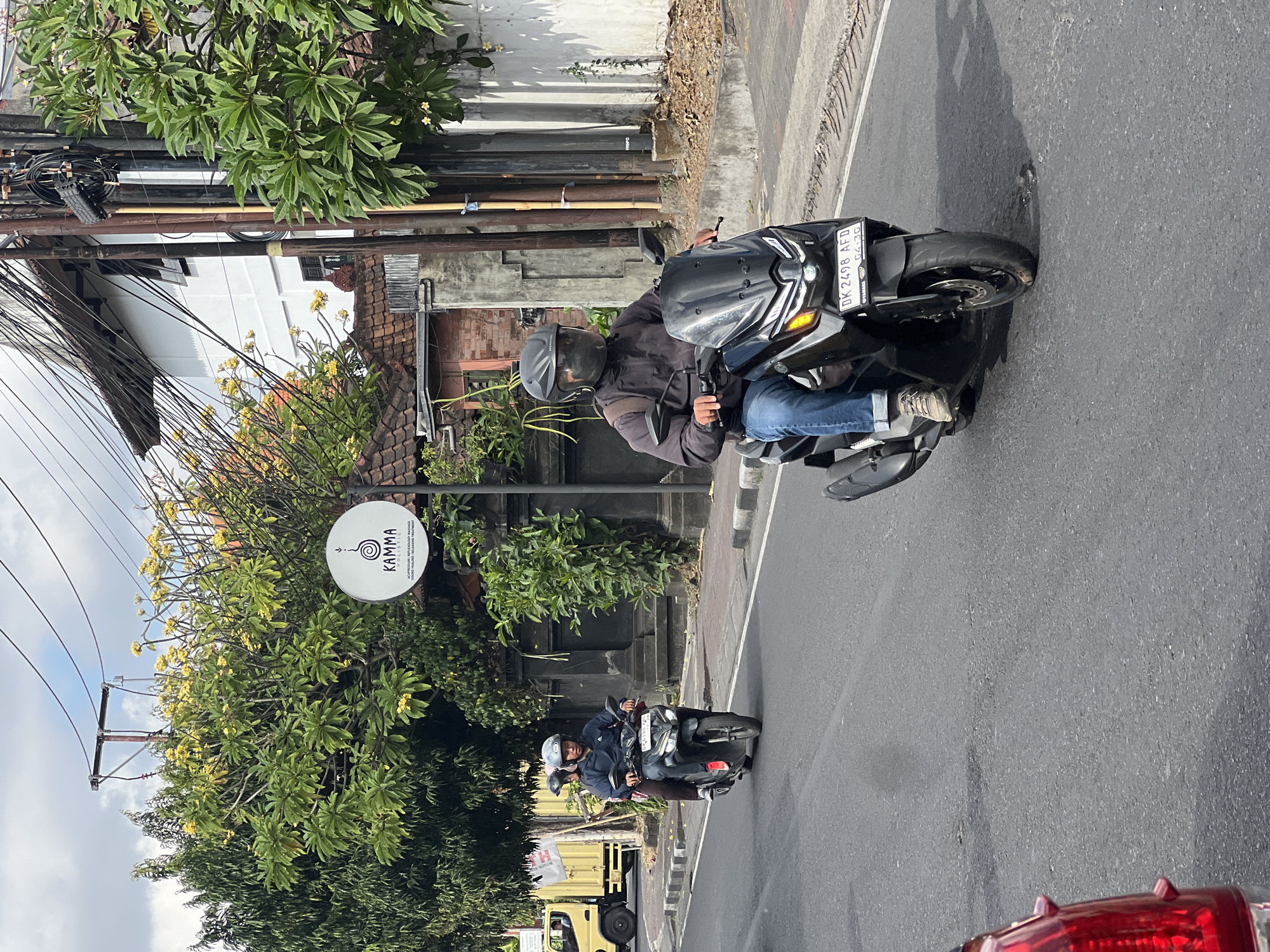 A man and woman ride a black scooter on a street in Denpasar, Indonesia. The man is driving, wearing a black jacket, blue jeans, a grey helmet with a tinted visor, and light brown shoes. The woman is the passenger, wearing a dark jacket, dark pants, and a black helmet. The scooter has a license plate that reads "DK 2498 AFD". Behind them, another scooter with two people is visible. In the background, trees with yellow flowers, a white building with a tiled roof, and overhead power lines are present. A sign with the text "KAMMA HOLISTIC" is also visible. The street is paved with asphalt, and there are white and black striped road markings. The sky is partly cloudy.